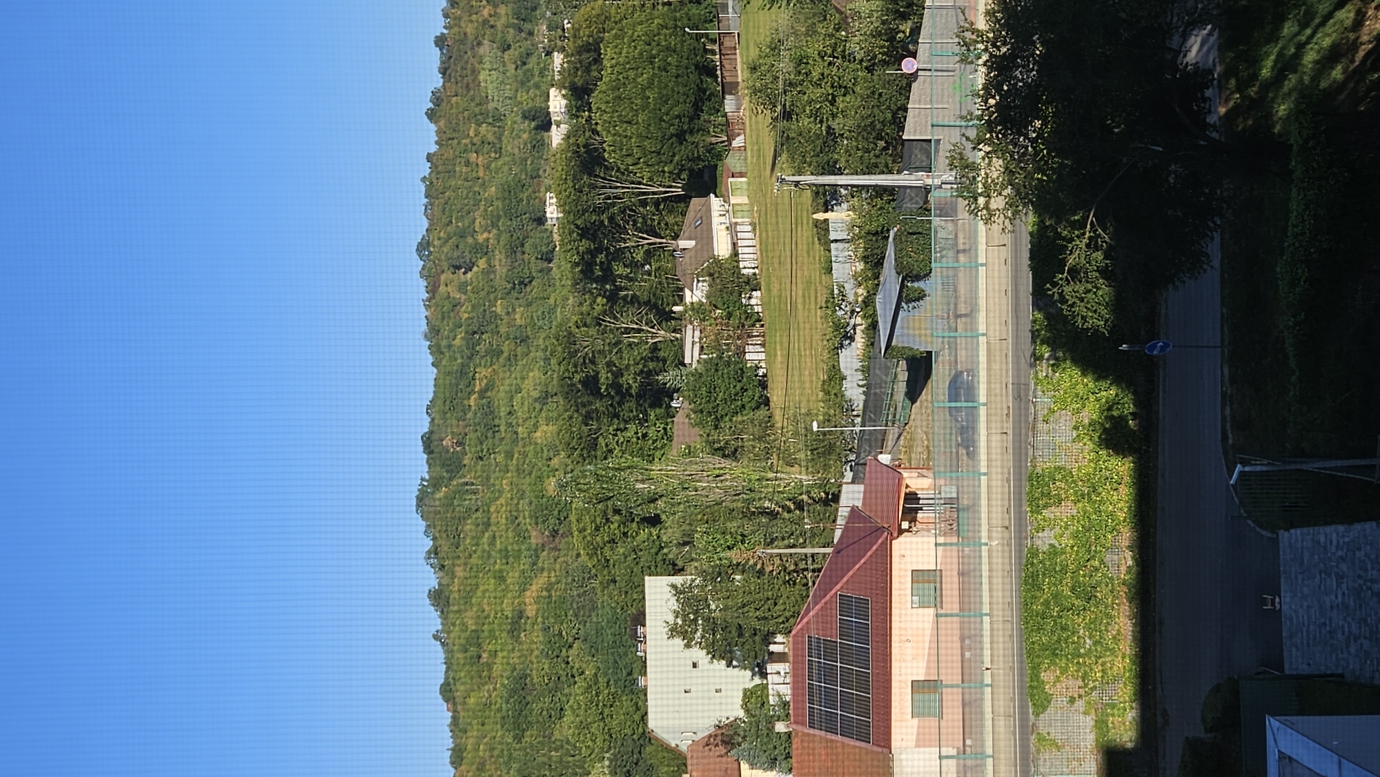 This is a telephoto vertical shot of a suburban landscape on a clear day, featuring a residential area nestled against a wooded hillside. In the foreground, a light-colored building with a red-tiled roof adorned with solar panels is visible. Adjacent to it is a road with a dark surface, bordered by a wall covered in lush green ivy. Further back, a row of houses with varied roofing and architectural styles are spread across a green, grassy area. Some of these houses are partially obscured by mature trees. In the distance, the hillside is densely covered with deciduous trees, displaying varying shades of green, indicating a summer or early autumn season. The sky above is a uniform, bright blue. A light pole with a blue circular sign is positioned on the right side of the road. The scene is calm and appears to be a residential area within or on the outskirts of Prešov, Slovakia, possibly near the Šariš neighborhood known for its hills and greenery. There are no people visible in the image.
