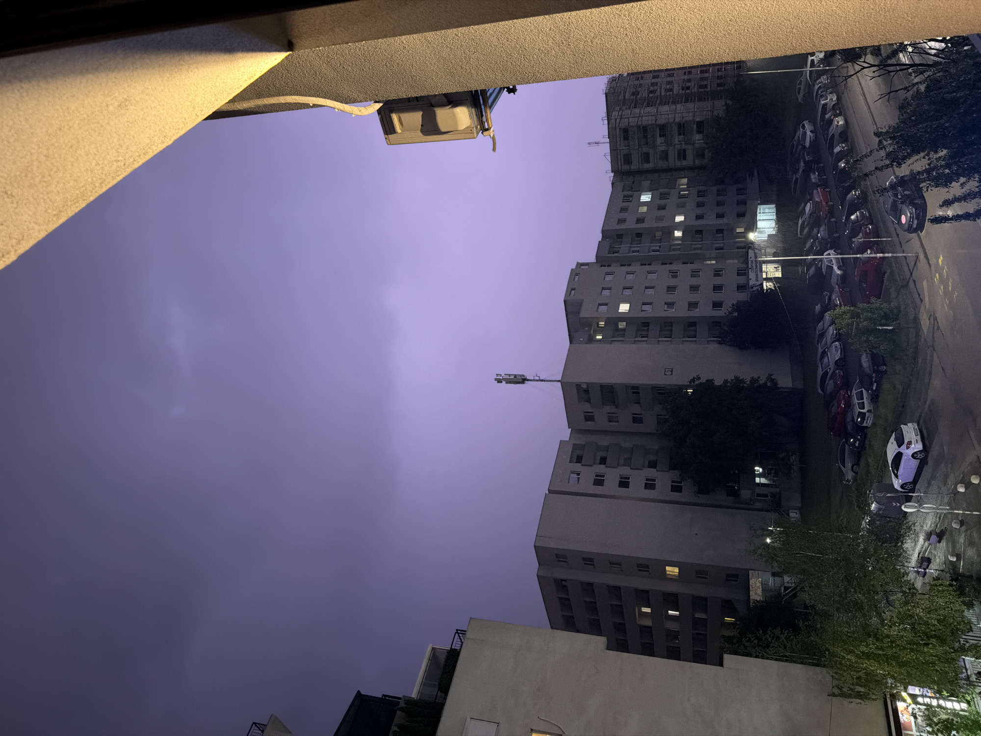 The image depicts an urban scene at twilight, likely in Sofia, Bulgaria, as indicated by the context of the provided information. The sky is a deep purple, suggesting a storm or the fading light of day, with faint cloud formations visible. In the foreground, a portion of a beige building facade with textured concrete and a bright yellow light source on its right side frames the view.
Dominating the mid-ground are several multi-story apartment buildings, appearing to be of Soviet-era architecture, characterized by their uniform window arrangements and grey concrete exteriors. Some windows are illuminated, indicating occupancy. One building on the right has visible scaffolding, suggesting ongoing construction or renovation.
Below the buildings, a street and a parking lot are visible. Numerous cars are parked in rows. Among them, a white hatchback is prominent in the lower left quadrant. Trees and greenery are scattered throughout the scene, some appearing lush and full, others more sparse. A telecommunications antenna is mounted on top of one of the apartment buildings. The overall atmosphere is calm, despite the potentially stormy sky, with no visible human activity.