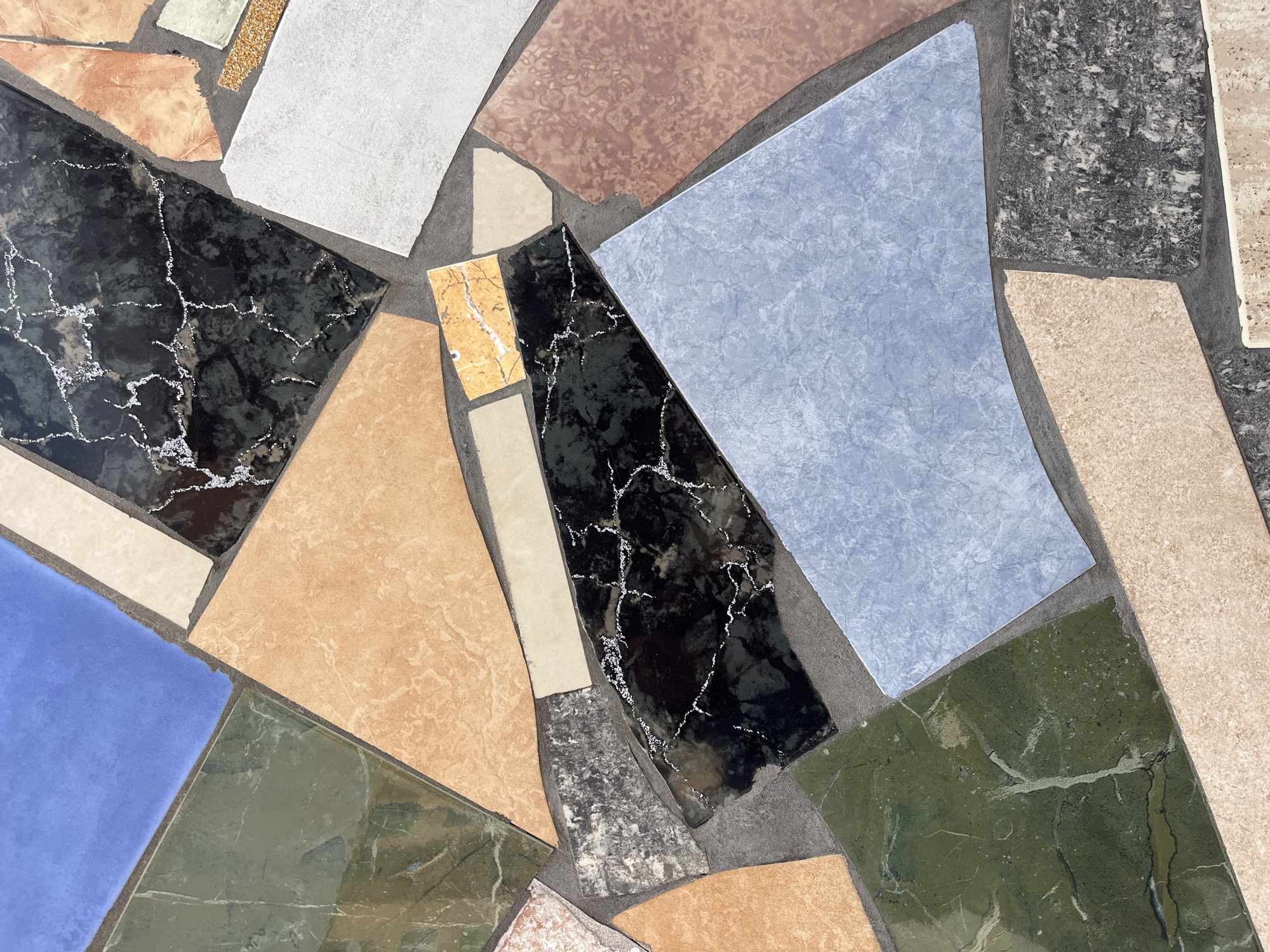 This image presents a close-up, top-down view of an abstract mosaic surface, located in the city of Varna, Bulgaria. The mosaic is composed of numerous irregularly shaped and sized tesserae set into a dark gray grout. The individual pieces consist of various materials, including ceramic tiles and what appear to be polished and unpolished natural stone or marble fragments.
The color palette is diverse, featuring a large, solid royal blue piece, several irregular dark green pieces with white veining, and multiple light orange/tan pieces, some plain and some with subtle marbling. Prominently featured are pieces of black material with distinct white, web-like veining, resembling black marble. Other components include a light gray textured piece, a reddish-brown textured piece, and various beige and off-white fragments, some plain and some with speckled or marbled patterns. One light blue, textured tile is visible towards the bottom-center. The edges of the tesserae are uneven, consistent with broken or hand-cut pieces, and the grout lines vary in thickness. No human figures, objects beyond the mosaic components, or specific actions are depicted. The scene is static, focusing solely on the textural and chromatic patterns of the mosaic.