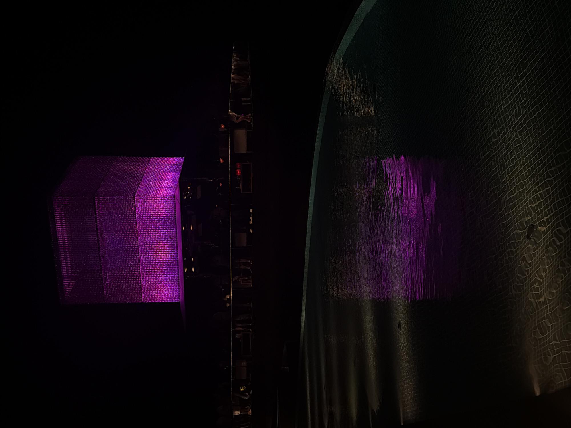 At night in Kuta Selatan, Indonesia, a purple illuminated, cuboid structure stands to the left of a dark, vertically oriented architectural structure. Further to the right is a dark, textured surface, possibly a wall or part of a building, also reflecting the purple light. The surroundings are largely obscured by darkness.