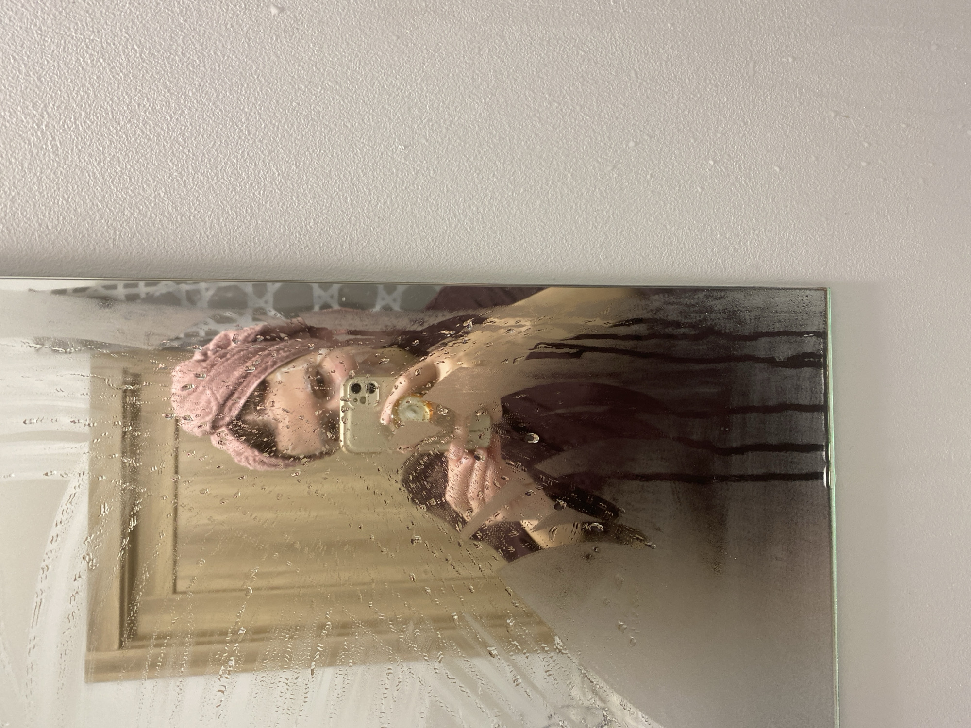A selfie of a woman is reflected in a steamed-up mirror. She is wearing a pink towel on her head and holding a mobile phone in her right hand. The background visible in the mirror is a wall with a light-colored frame. The scene appears to be inside a bathroom. There is no indication of the location being in Yekaterinburg, Russia.
