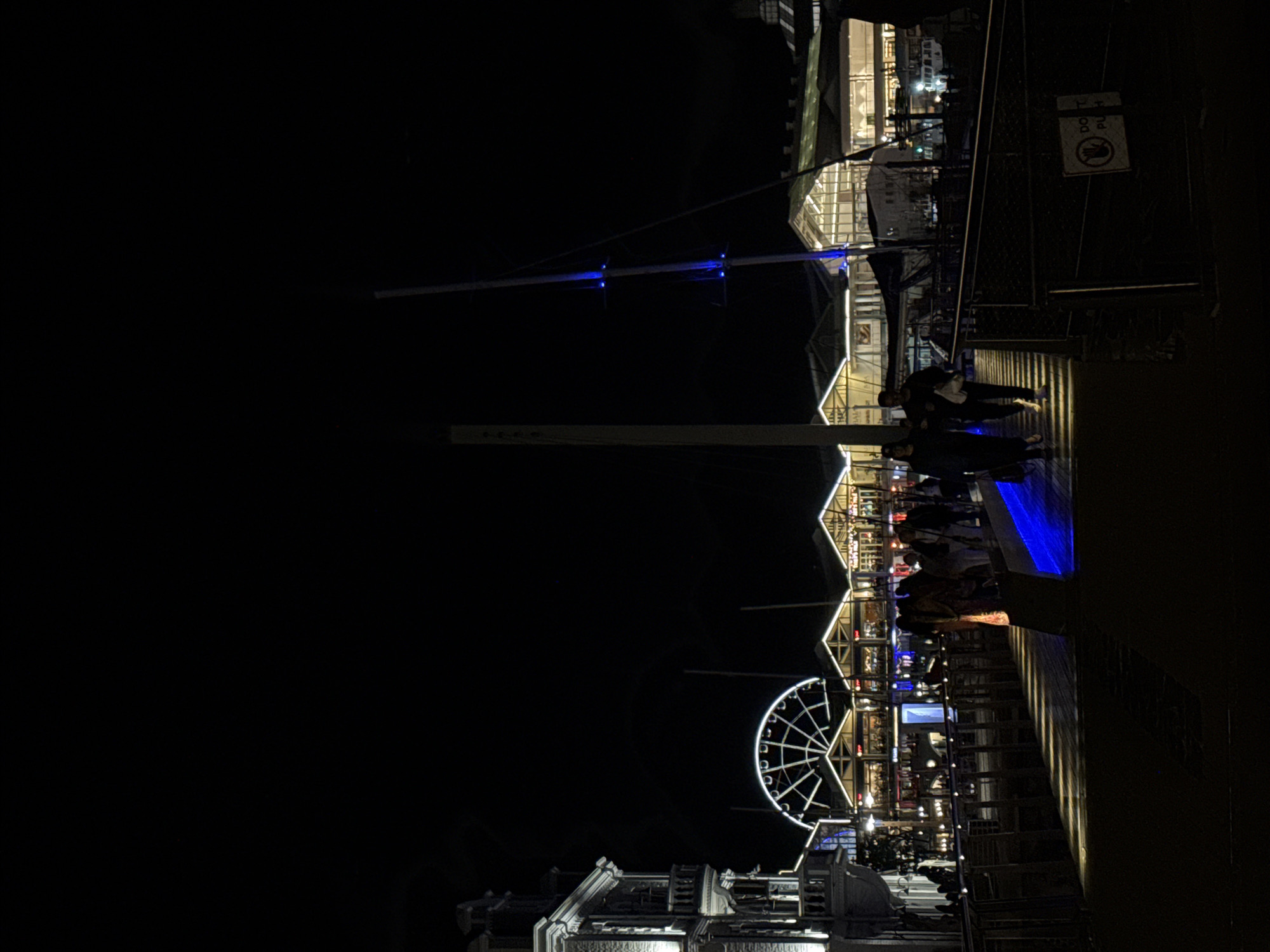 The image shows the Victoria & Alfred Waterfront in Cape Town, South Africa at night. A Ferris wheel, illuminated with white lights, stands towards the bottom left. Structures with angled roofs and numerous windows are visible along what appears to be a pier or walkway. People can be seen walking along the pier. A tall pole with blue lights is to the top left of the image. A sign with the words "DO NOT PLAY" and an icon of a person falling over is on the right side of the image. The environment is generally dark with the scene being illuminated by lights from the waterfront area.