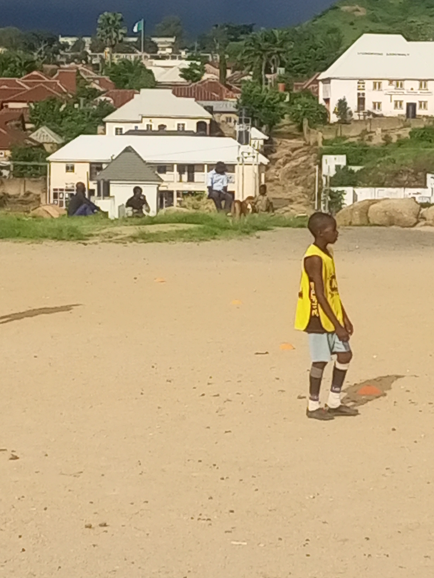 In the city of Jos, Nigeria, a young boy wearing a yellow jersey and blue shorts stands on a dirt field, looking to his right. He is wearing dark socks pulled up to his knees and brown shoes. Scattered across the field are several orange cones. In the background, a cluster of buildings with red tiled roofs and white walls are visible, some appearing to be residential homes. Beyond the buildings, a green, hilly landscape rises, topped with trees. A Nigerian flag flies from a pole in the distance. A group of people are seated on a grassy area in front of some of the buildings. The scene appears to be a relatively calm, outdoor setting in a suburban area of Jos.