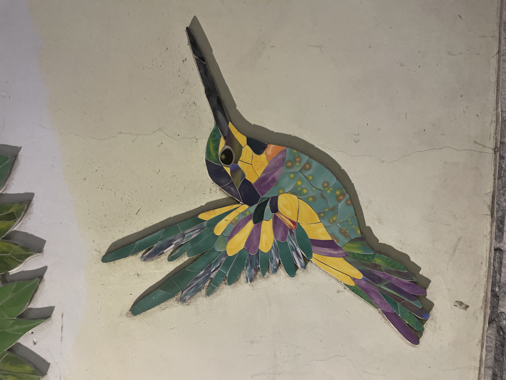 A mosaic of a hummingbird in flight is mounted on a light-colored, textured wall. The hummingbird is depicted with wings outstretched, composed of numerous small, irregularly shaped tiles in shades of green, yellow, purple, and blue. Its body features teal tiles with small orange speckles. The bird's beak is long and pointed, rendered in dark gray tiles. The wall has visible cracks and some darker spots, suggesting an aged or outdoor setting. Above the hummingbird, a portion of another mosaic, appearing to be green leaves, is visible. The bottom of the image shows a textured gray band, possibly a baseboard or the edge of another decorative element. The lighting creates a shadow of the hummingbird on the wall, extending to its right.