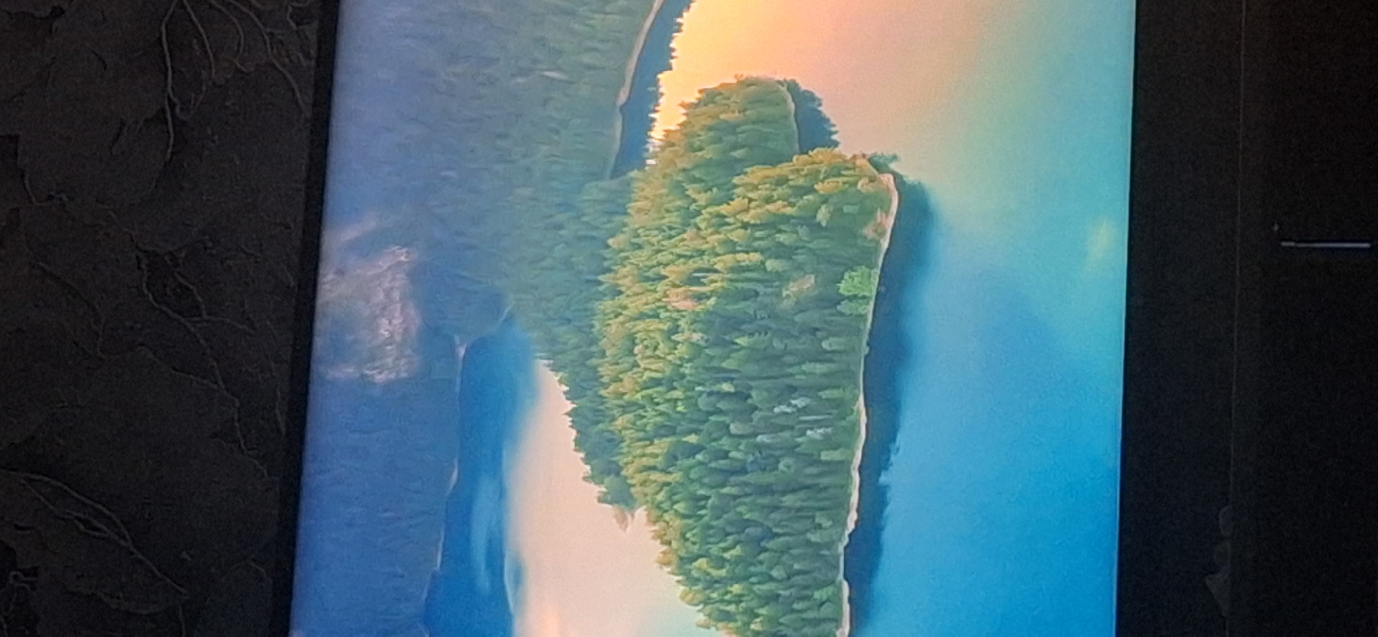 The image displays an aerial view of a densely forested island surrounded by clear blue water. The island is covered in a multitude of evergreen trees, creating a thick canopy. The water is calm and reflects the sky, which appears to have some light clouds or atmospheric haze. The lighting suggests it is either early morning or late afternoon, with a warm glow visible on the right side of the image, possibly indicating a sunrise or sunset. The scene is natural and serene. There is no visible indication of the city of Bila Tserkva, Ukraine.