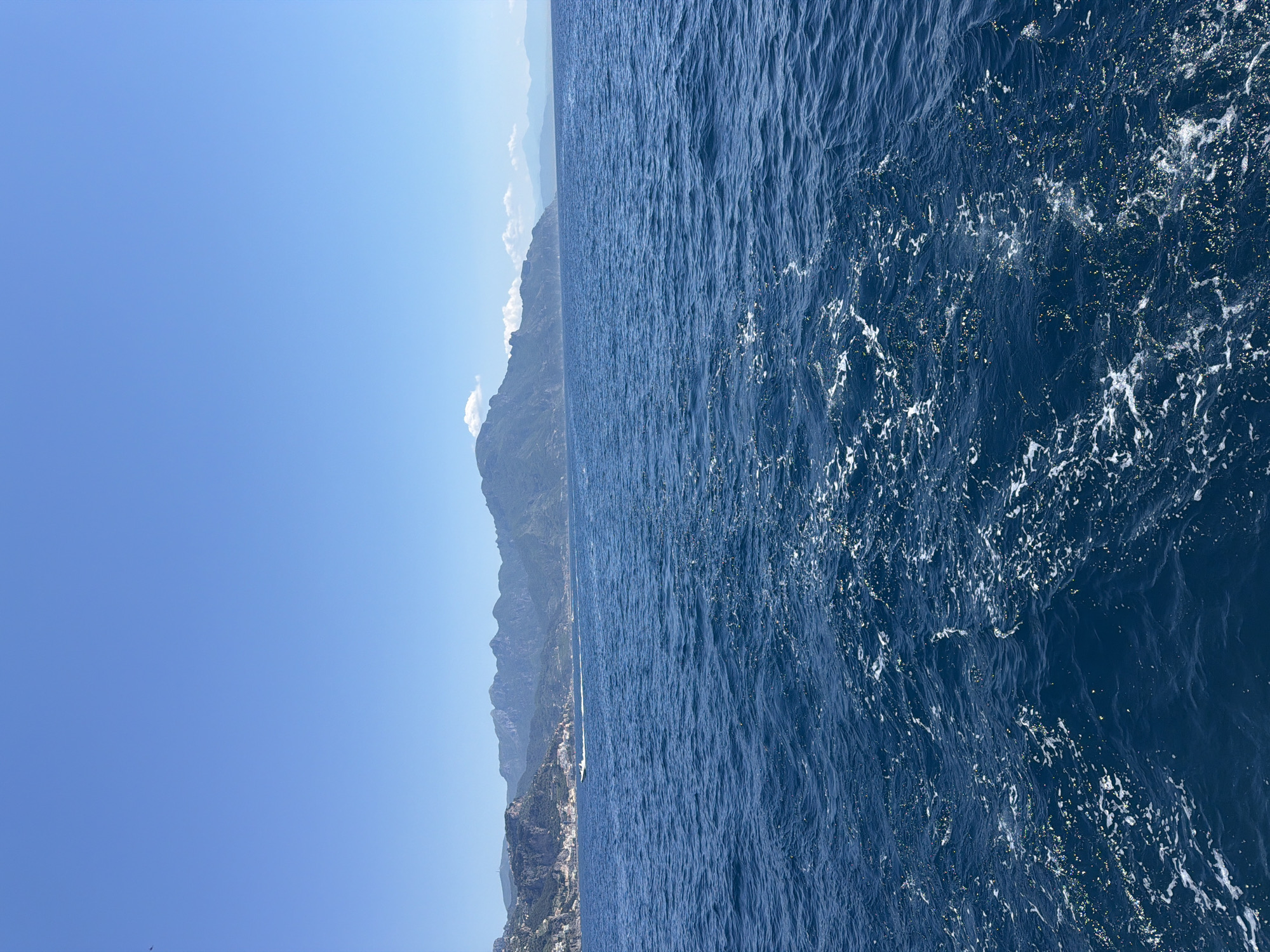 The image displays a view of the Amalfi Coast in Conca dei Marini, Italy. On the left, a clear, blue sky fades into the horizon. Adjacent to the sky, a mountainous coastline stretches along the frame. The base of the mountains is interspersed with structures, likely part of the town of Conca dei Marini. To the right, the deep blue water of the Mediterranean Sea fills the frame, with visible ripples and white foam patterns, indicating movement or waves.