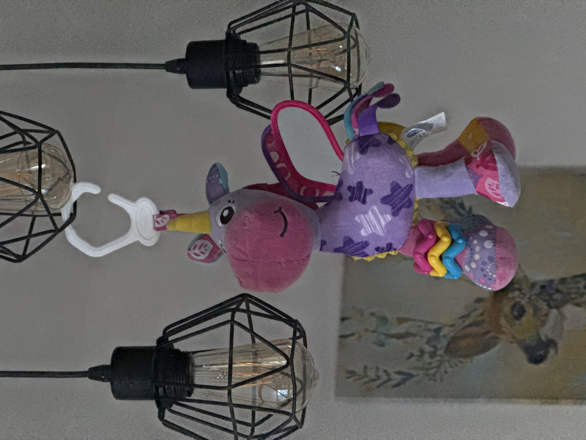A plush toy unicorn, primarily purple and pink with yellow accents, hangs from a white clip attached to a black wire cage lamp fixture. The unicorn has a smiling face, a yellow horn, pink ears with leaf designs, and colorful hooves. It has iridescent wings. Behind the unicorn, a framed artwork depicting a deer with foliage is visible. Two other black wire cage lamp fixtures are present, one on the left and one on the right, each containing a lit Edison-style bulb. The background is a plain, light-colored wall. The scene appears to be indoors, possibly in a residential setting. There is no indication of Bratislava, Slovakia, in the image.