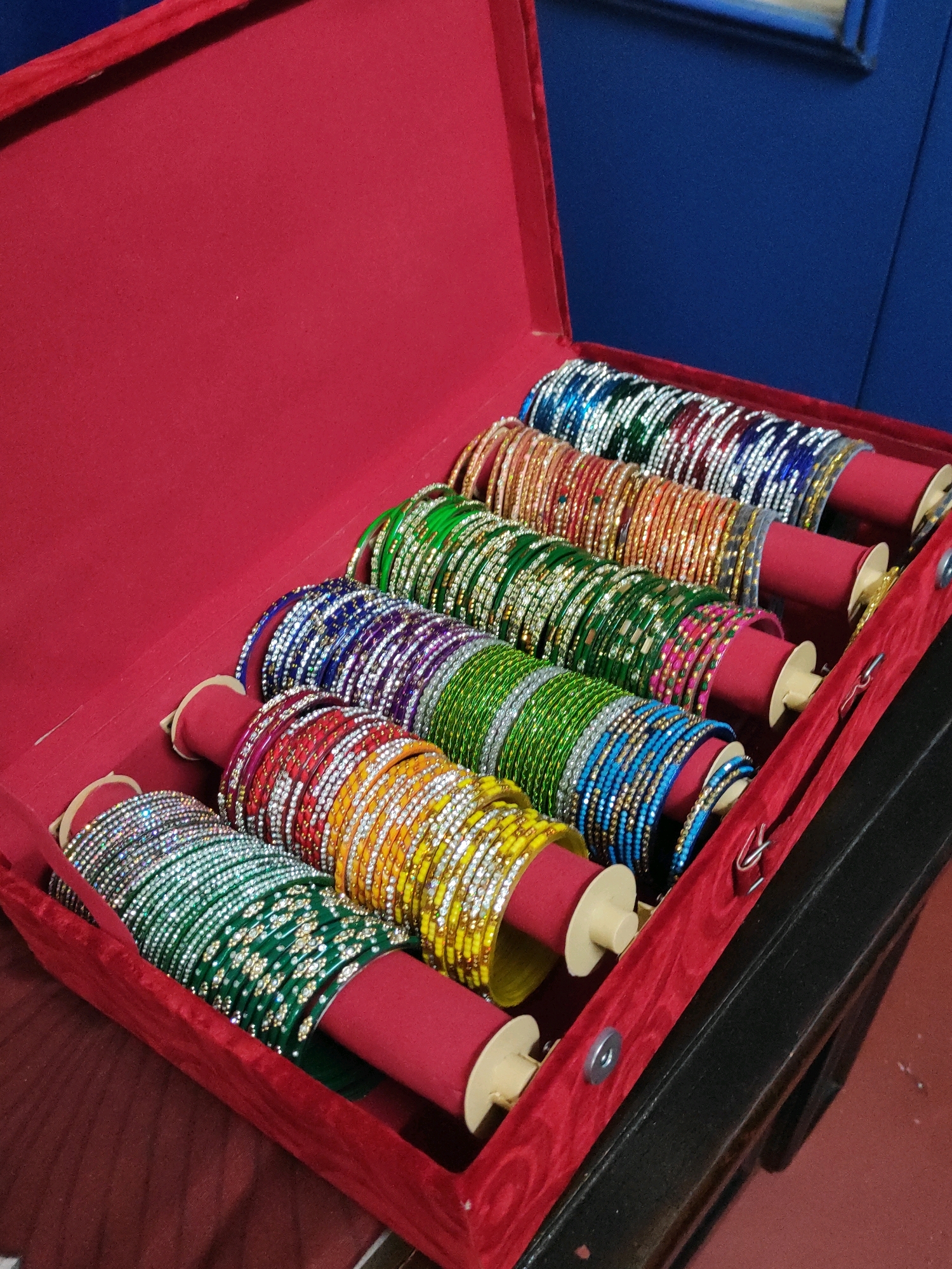 The image shows a red velvet jewelry box filled with rows of colorful bangles. The bangles are adorned with small, sparkling stones. They are arranged on individual red cylindrical stands within the box, which is open. The background is a deep blue. This image was taken in the city of Hyderabad, India.