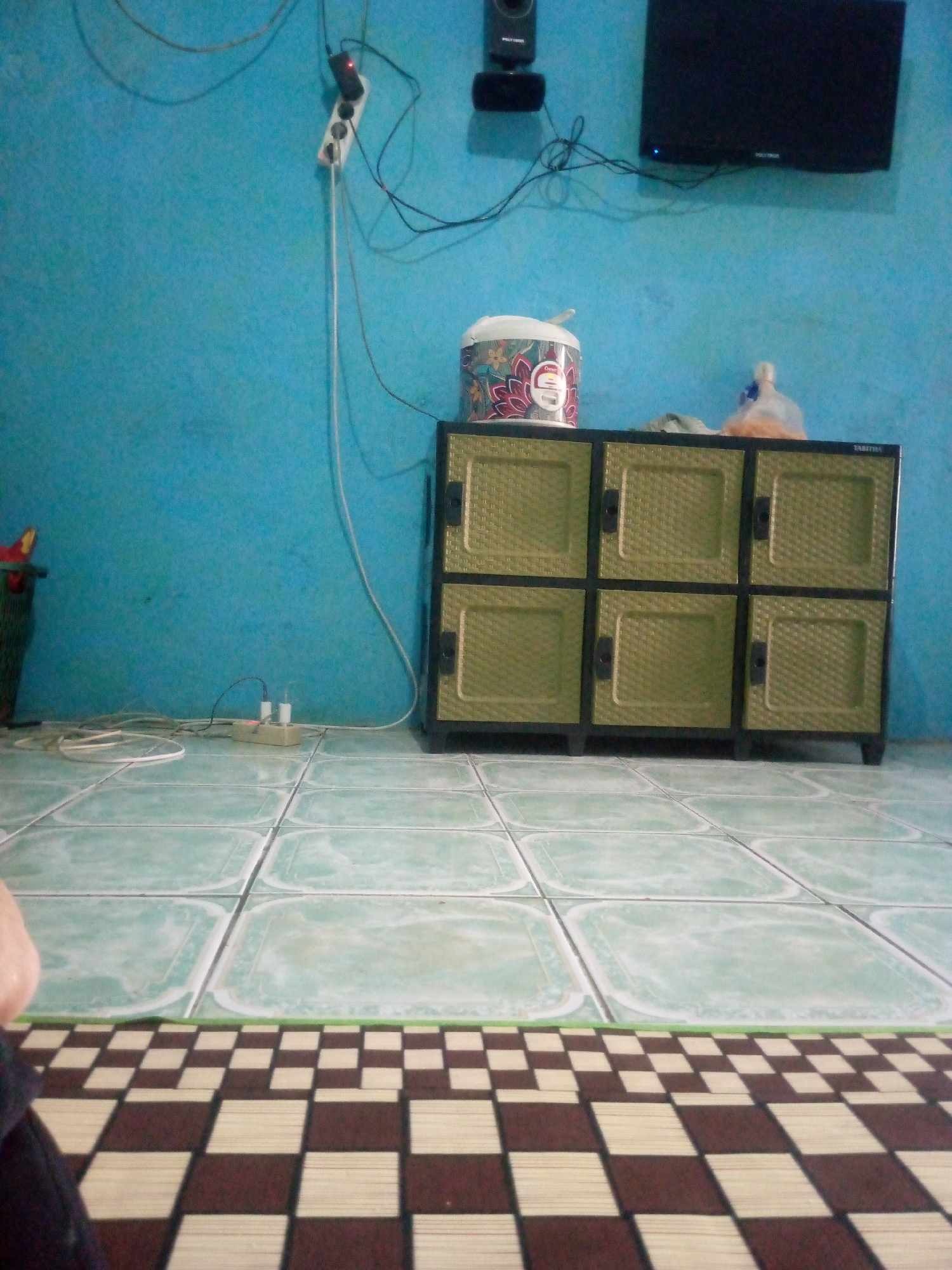 The image depicts an interior scene in Giriasih, Indonesia, likely a residential room. The wall is painted a vibrant blue and is adorned with electrical wiring, a power strip with two plugs, and a television mounted on the upper right. Below the television, a woven plastic cabinet with six doors is positioned against the wall. On top of the cabinet, a white rice cooker with a floral pattern is visible, along with a plastic bag containing items. The floor is tiled with a light green pattern, and a checkered mat lies in the foreground, partially obscuring a person's leg on the left side of the frame. To the far left, a green trash can is partially visible. The overall impression is of a utilitarian living space.