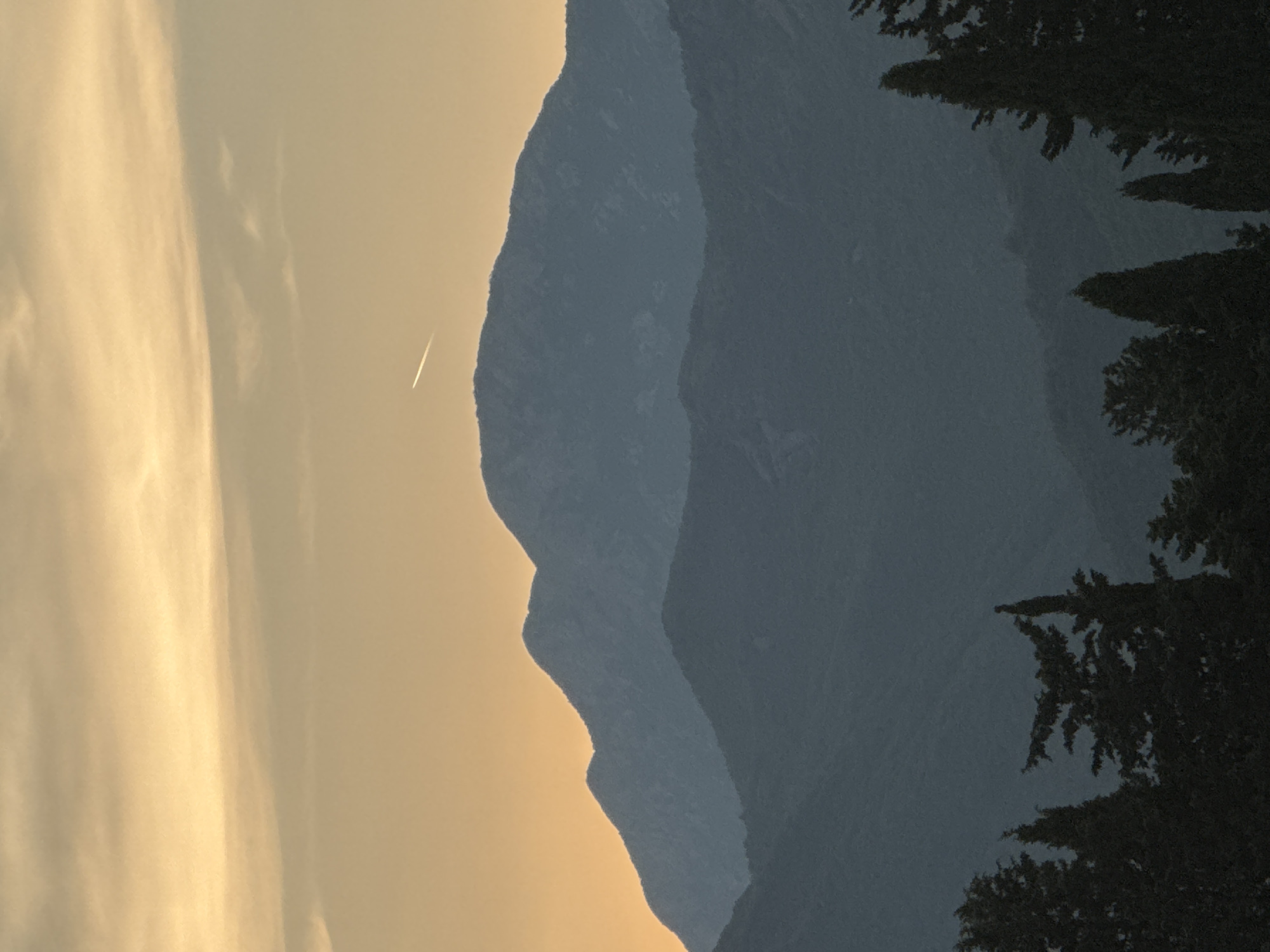 The image displays a silhouetted landscape with layered mountain ranges fading into the distance. In the foreground, dark, jagged shapes of pine trees are visible on the right side of the frame. The sky is a gradient of warm, golden hues on the left, transitioning to a dusky blue behind the mountains. A thin contrail from an airplane is visible against the lighter sky, indicating activity at a high altitude. The overall impression is one of a serene, natural setting, possibly at dusk or dawn, with the soft light creating a sense of depth and tranquility. There is no visible text in the image.