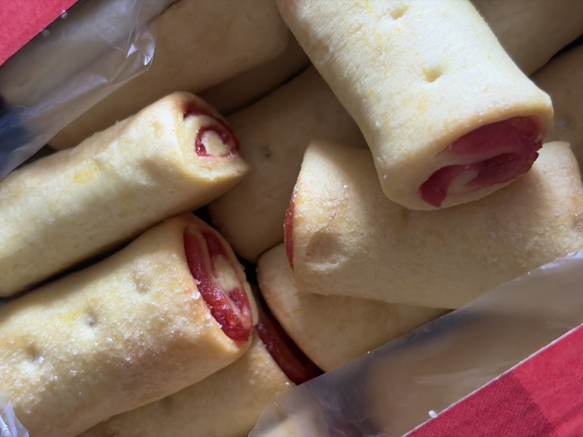 The image shows a close-up of a box of pastries. The pastries are cylindrical, with a light-colored dough exterior and a red jam filling visible at the ends. The pastries are coated with sugar crystals. The background includes a clear plastic wrapper and a red box. There is no information in the image to indicate the location of Saint Petersburg, Russia.