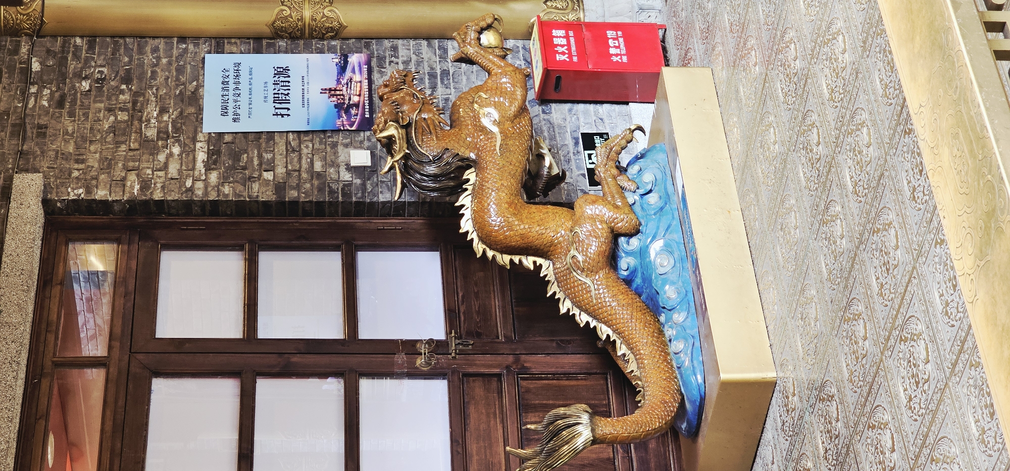 The image displays a large, ornate golden dragon sculpture positioned in front of a wooden door and a brick wall. The dragon is depicted in a dynamic pose, appearing to leap forward from a blue, wavy base that simulates water. It has a long, scaly body with golden spines along its back and tail, and sharp claws. The dragon's head is detailed with horns, a mane, and an open mouth. To the right of the sculpture, a bright red fire extinguisher box is visible, bearing Chinese characters and English text indicating "FIRE EXTINGUISHER BOX" and "FIRE TELEPHONE 119". A poster with Chinese text is mounted on the brick wall behind the dragon and the extinguisher box. The floor is covered with intricately patterned tiles. The overall setting suggests an indoor or semi-outdoor public space, possibly a historical or cultural venue. The presence of a sign with what appears to be official regulations or announcements, combined with the dragon sculpture, might indicate a location in Yuzhong District, China, known for its blend of historical architecture and modern development.