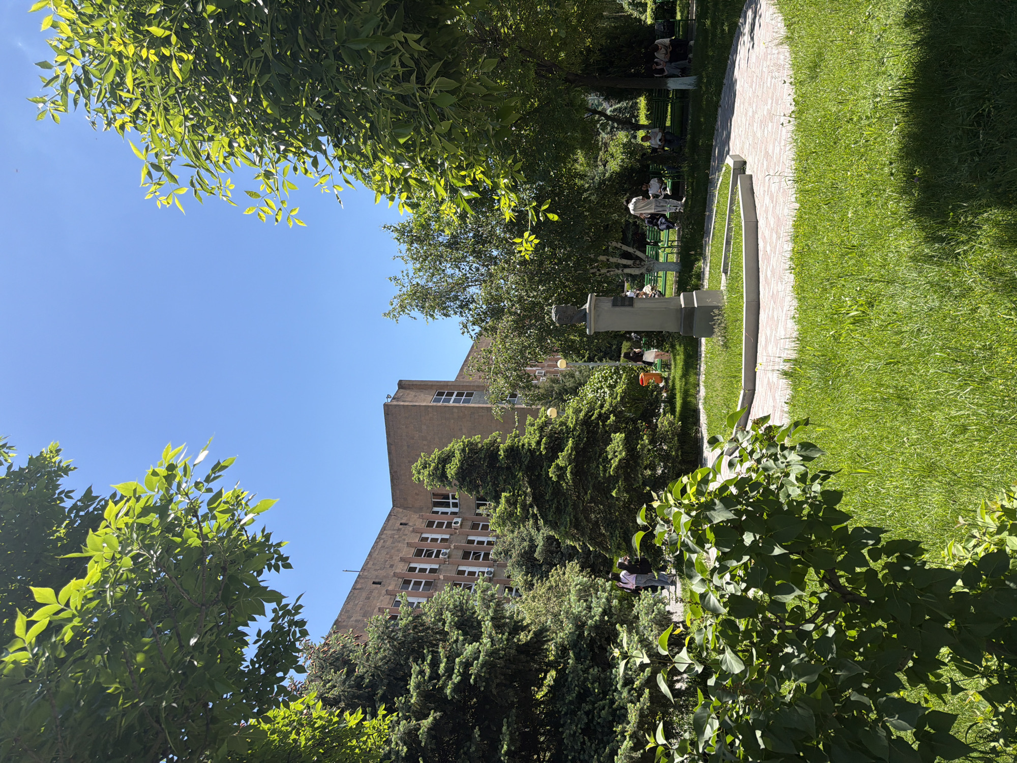 The image shows a park in Yerevan, Armenia. There's a building in the background and a stone path running along the right side of the image. Grass is visible on either side of the path. Several people are seated on benches in the background. A stone monument stands next to the path. Trees and other greenery frame the scene. The sky is clear and blue.