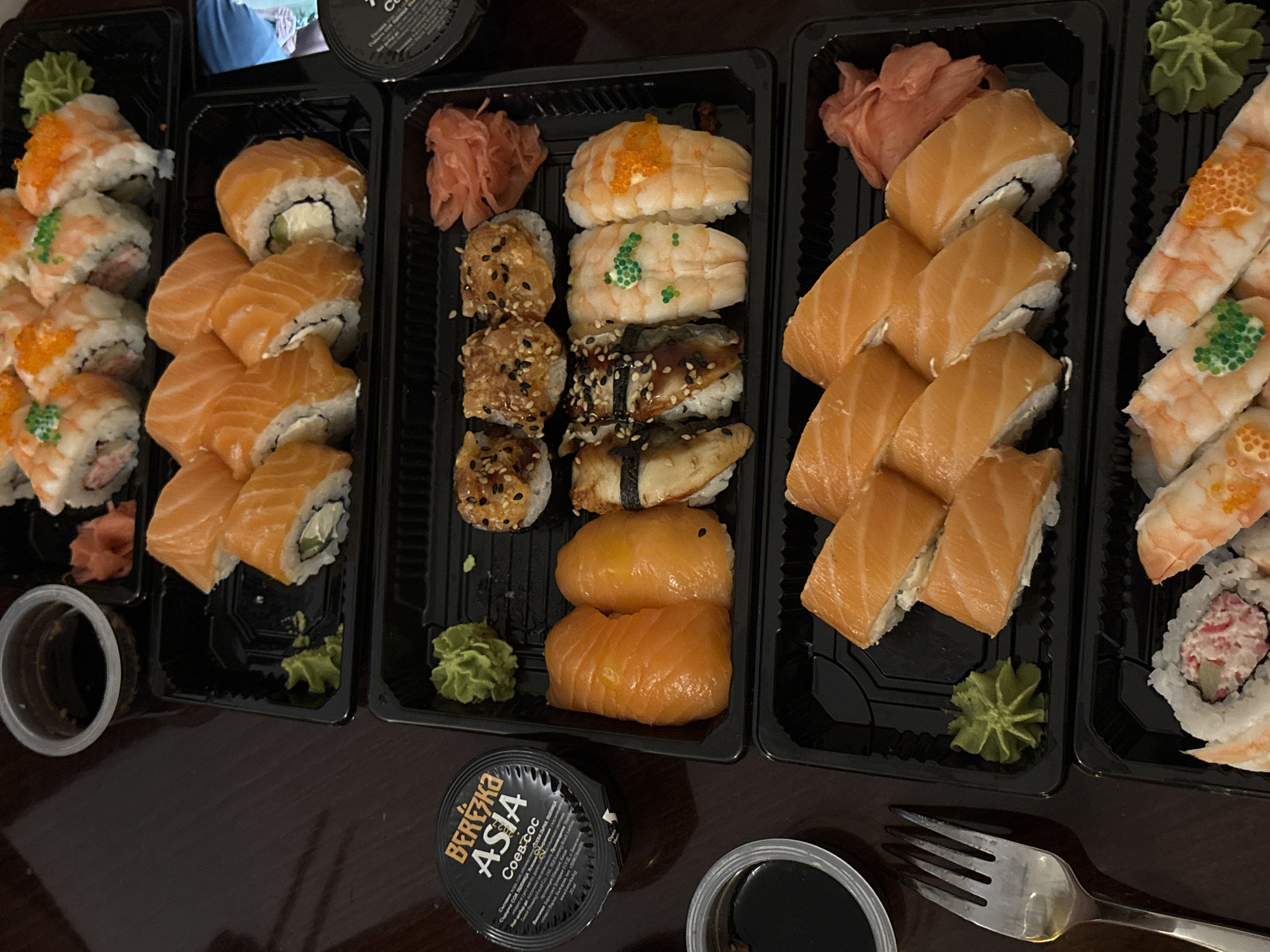 A close-up, overhead shot displays a dark brown, wood-grain table surface populated with an assortment of sushi dishes. In the upper left corner, a small, clear plastic container holds dark soy sauce. Adjacent to it, a black plastic container, labeled in Russian with "Березка ASIA" and "Соев-сос", sits open. The bulk of the image is filled with three black plastic trays containing various sushi rolls and nigiri. The top tray features salmon nigiri and rolls with salmon and cream cheese. The middle tray holds eel nigiri and what appear to be shrimp nigiri, some adorned with green and orange tobiko or masago. The bottom tray presents more salmon rolls. Pickled ginger is placed in the upper right corners of the middle and bottom trays. A silver fork rests on the table to the left of the bottom sushi tray. The scene is devoid of people, indicating an at-home or takeout setting. There is no discernible information or visual cues within the image to suggest a specific location in Varna, Bulgaria.