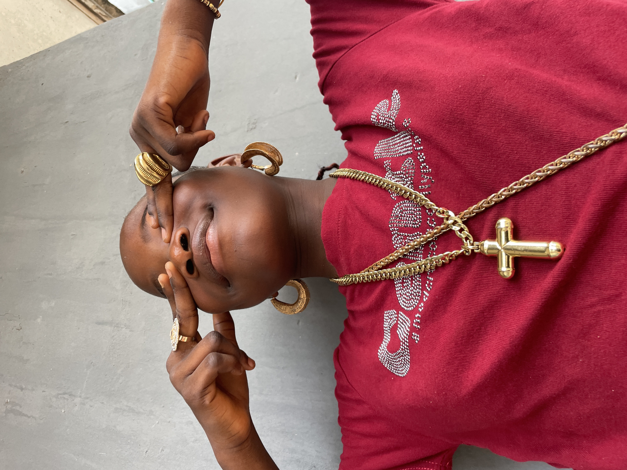 The image presents an intimate, angled portrait of a young Black individual from Badagry, Nigeria, lying on a plain, light grey concrete surface. The person's head is positioned towards the left side of the frame, with hands gently brought up to their face in a thoughtful or contemplative pose. One finger lightly touches the nose, while another hand rests near the mouth and chin.
The individual is dressed in a red t-shirt, which features an intricate silver-white beaded or sequined design on the chest. While decorative, these designs do not form clearly legible text. A variety of prominent gold jewelry adorns the person: multiple gold rings are visible on their fingers, including a wide, textured ring on the index finger and a more ornate, possibly jeweled, ring on another. Large, thick gold hoop earrings are worn in both ears. A substantial gold chain necklace, featuring a prominent golden cross pendant, rests on the red shirt. A gold beaded bracelet is also visible on one wrist.
The subject's eyes are open, looking slightly upwards, conveying a composed and relaxed expression. The simple, uncluttered background keeps the focus on the individual and their accessories. The lighting indicates it is daytime, though no specific time or weather conditions can be determined from the image.