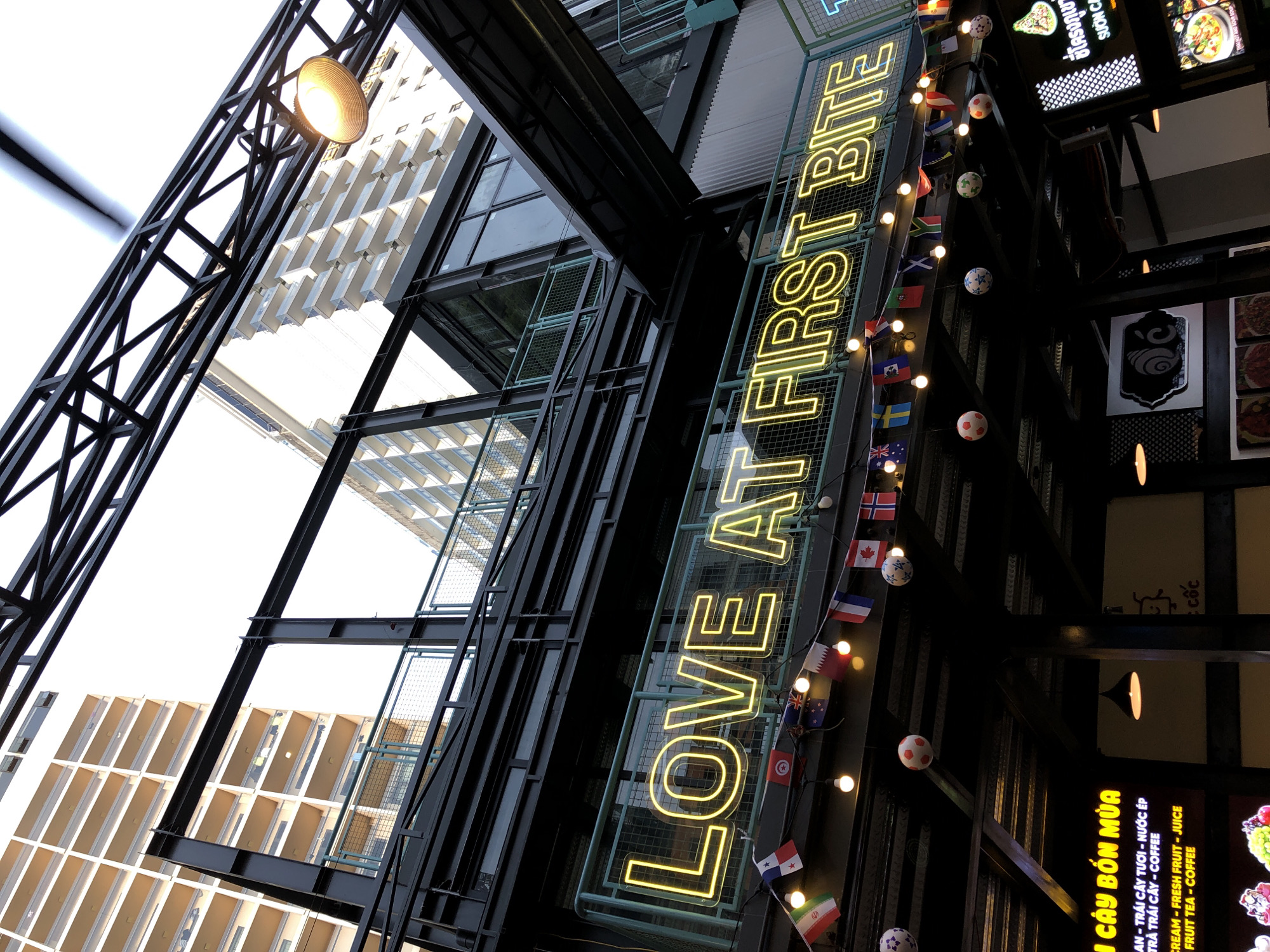 The image shows the exterior of a food establishment in Đà Nẵng, Vietnam. A prominent neon sign reads "LOVE AT FIRST BITE" in bright yellow. Below the sign, a string of small international flags and light bulbs is visible, along with some decorative soccer balls. The building features a modern, industrial architectural style with dark metal beams and structures, contrasting with the lighter colors of the buildings in the background, which appear to be multi-story residential or hotel complexes. The perspective is from below, looking upwards towards the sign and the architecture. Text in Vietnamese is visible on a sign in the lower left, advertising various food and drink items such as "TRAI CAY BON MUA" (four seasons fruit), "CREAM - FRESH FRUIT - JUICE," and "FRUIT TEA - COFFEE." Another sign in Thai script is visible in the lower right. The overall scene suggests an outdoor dining or market area, likely in an urban setting.