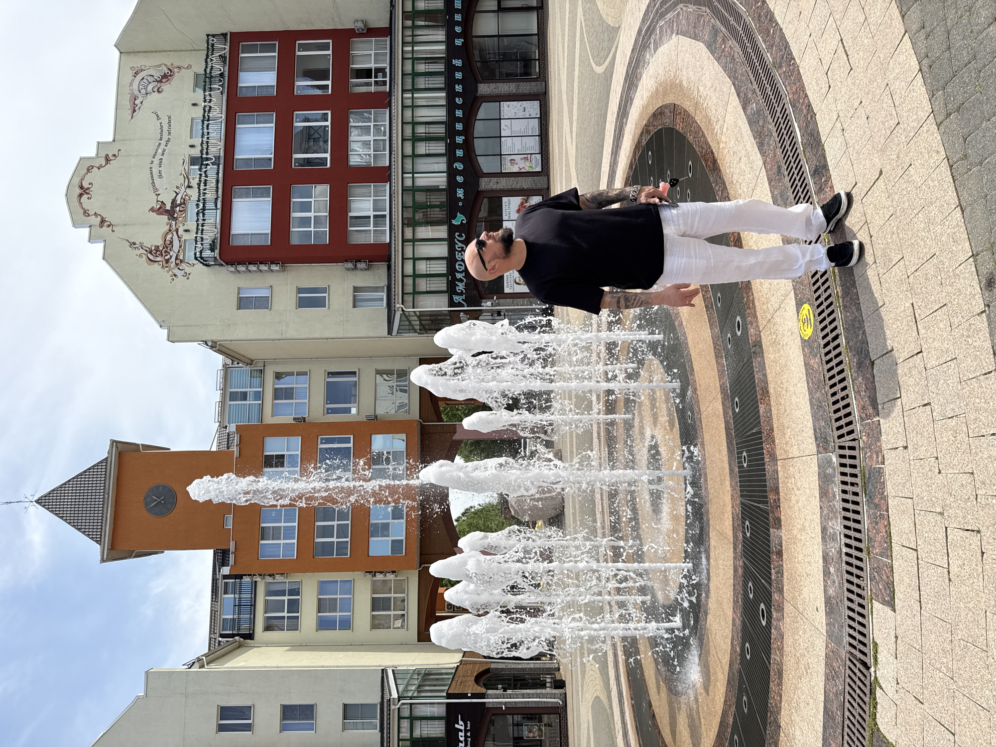 A man with a shaved head and tattoos stands in front of a fountain in a city square. He is wearing a black t-shirt, white linen pants, and black shoes. The fountain has multiple water jets and is surrounded by a circular patterned pavement. In the background, there are several buildings with distinct architectural styles, including one with an orange tower and a clock. A mural is visible on the facade of another building. The sky is clear with some clouds. The scene appears to be in Krasnodar, Russia, possibly on Theater Square, known for its fountains and surrounding cultural institutions.