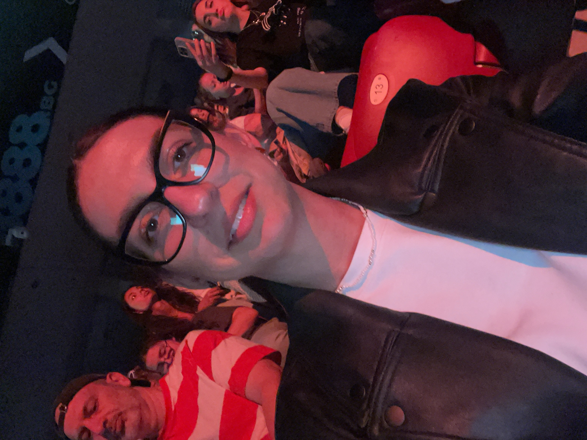 A selfie taken by a woman with glasses in a movie theatre. The theatre seats are red. People can be seen in the background. The image is taken in Sofia, Bulgaria, as evidenced by the "888 BG" markings in the top left of the photo.
