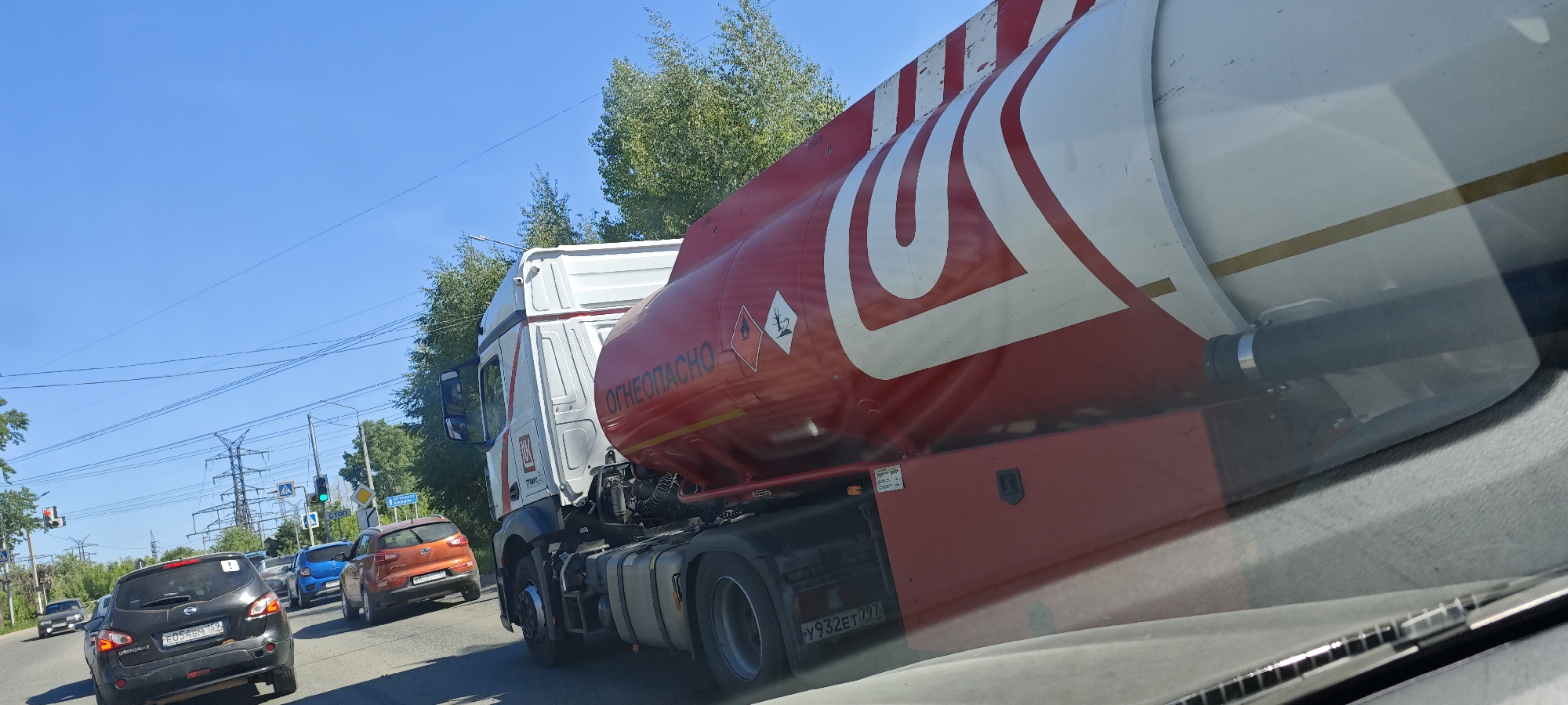 The image depicts a red and white tanker truck with the word "ОГНЕОПАСНО" (meaning "FLAMMABLE") written on it in Russian, likely carrying a flammable substance. The truck is in motion on a road with other vehicles, including a black Nissan Qashqai and an orange Kia. Power lines are visible in the background, as well as trees and clear blue sky. The photo seems to have been taken from inside a vehicle through the windshield, possibly on a road in Chaikovsky, Russia, given the license plates and the presence of Russian text.