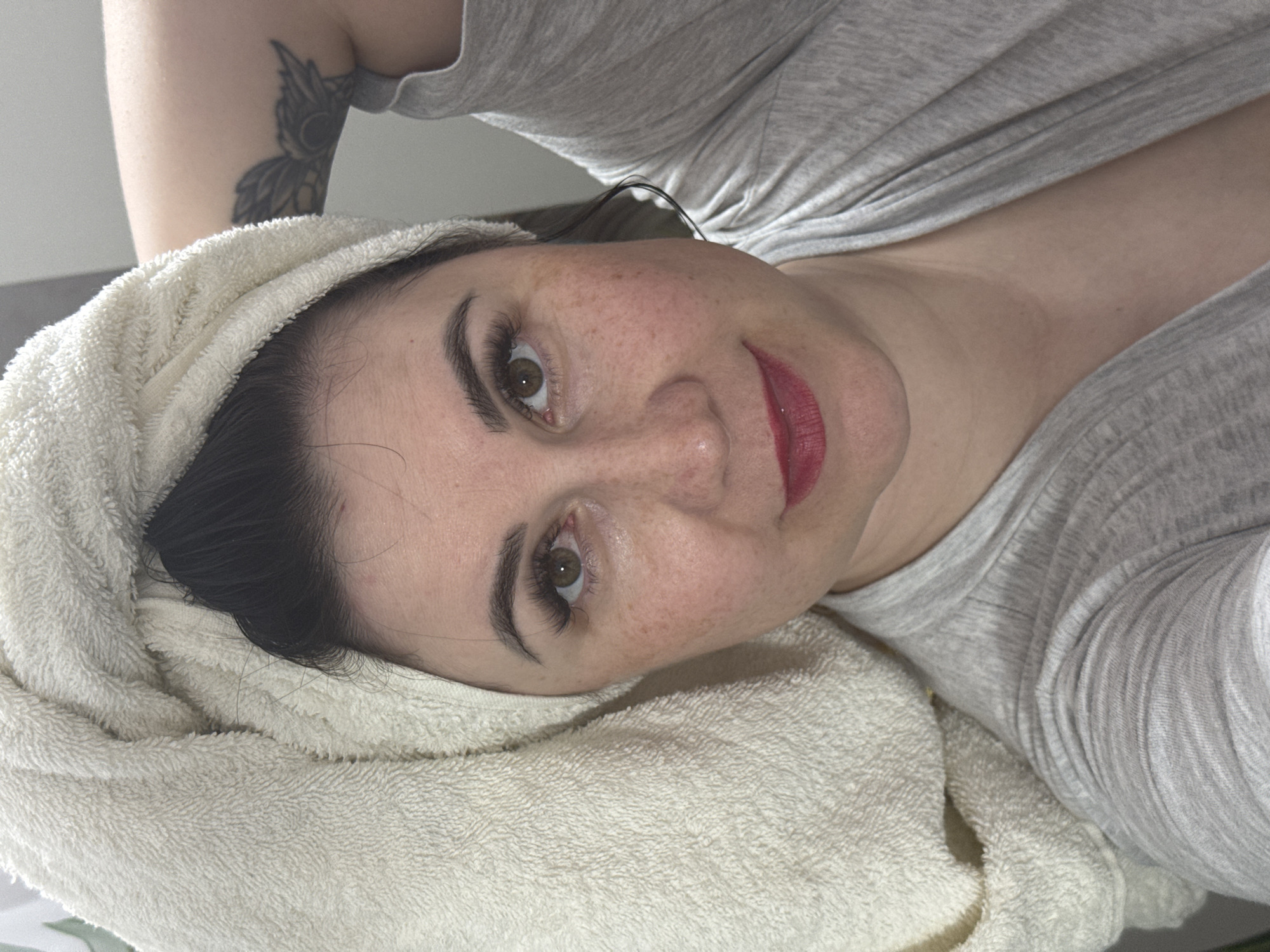A woman is lying down, facing the camera. She has fair skin, freckles, dark hair partially covered by a white towel wrapped around her head, and is wearing red lipstick. She has visible eyelashes and brown eyes. She is wearing a grey shirt. A tattoo is visible on her left arm. The background appears to be a plain wall. It is impossible to confirm the location of this image to Zav'yalovo, Russia.