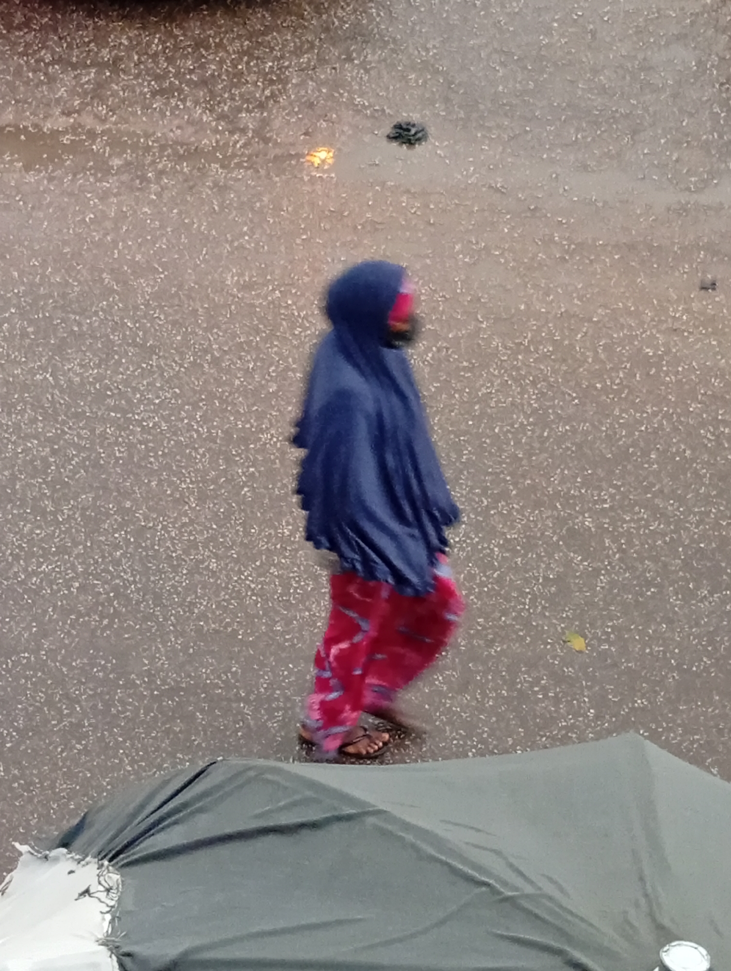 In Bukuru, Nigeria, a person is walking in heavy rain, dressed in a blue hooded garment and red patterned pants, with sandals on their feet. The individual is positioned in the center of the frame, while the ground is visibly wet from the rainfall. In the foreground, a portion of a green umbrella is visible.