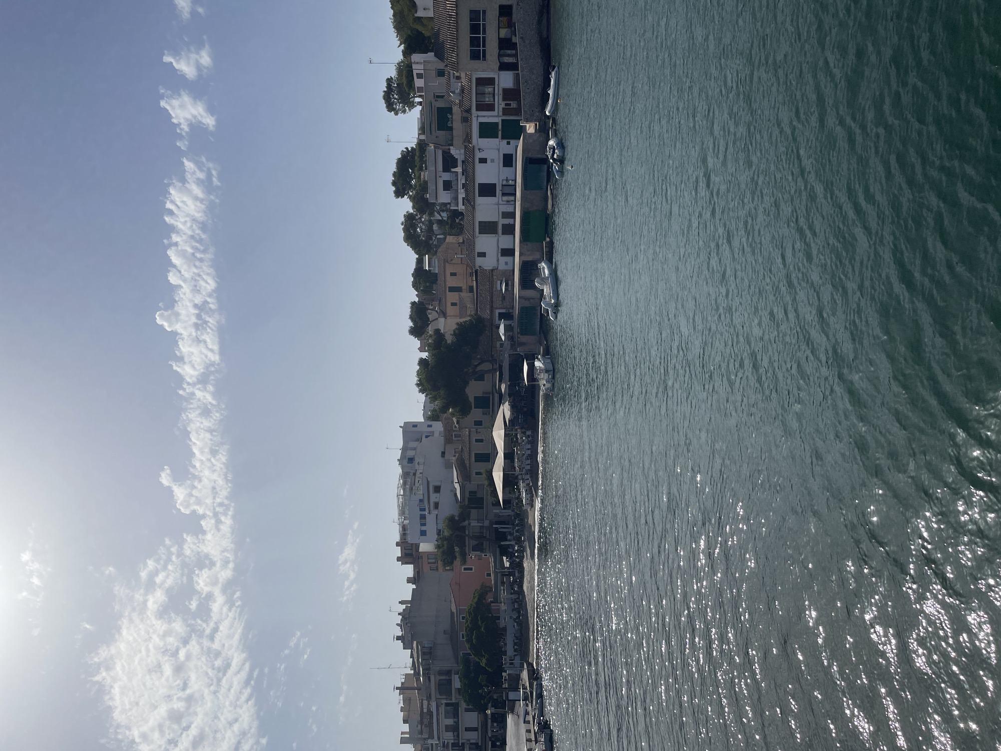 The image shows a waterfront scene in Portopetro, Spain. In the foreground, there is a body of dark blue water with sunlight glinting off its surface, creating a textured appearance. Along the middle ground, a row of buildings lines the shore. These buildings are varied in color and style, with some appearing to be residential and others commercial, possibly restaurants or shops, indicated by umbrellas and outdoor seating visible on a small beach area. Several small boats, primarily white dinghies and inflatable vessels, are moored in the water near the buildings, some of which have green boathouse doors. In the background, more buildings rise up, with some trees interspersed among them. The sky above is a pale blue with scattered white clouds, and the sun appears to be high in the sky, casting bright light. The overall scene depicts a tranquil coastal town with a harbor.