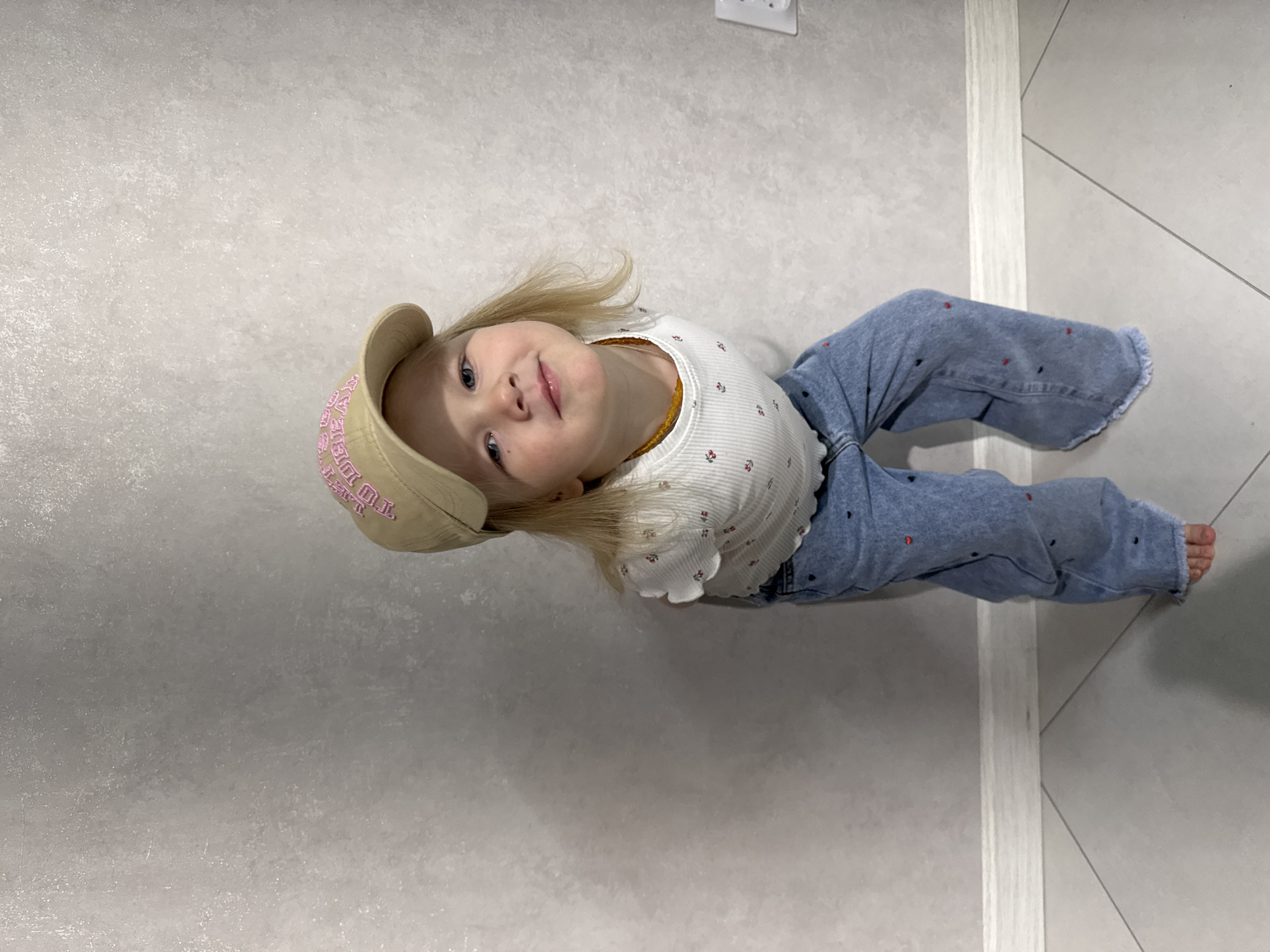 A young girl with blonde hair and blue eyes is lying on a gray and white tiled floor. She is wearing a beige baseball cap with pink lettering, a white long-sleeved shirt with small red floral prints, and blue jeans with small red and black heart-shaped patterns. Her feet are bare. The location appears to be indoors, possibly a home. The lighting is bright and even.