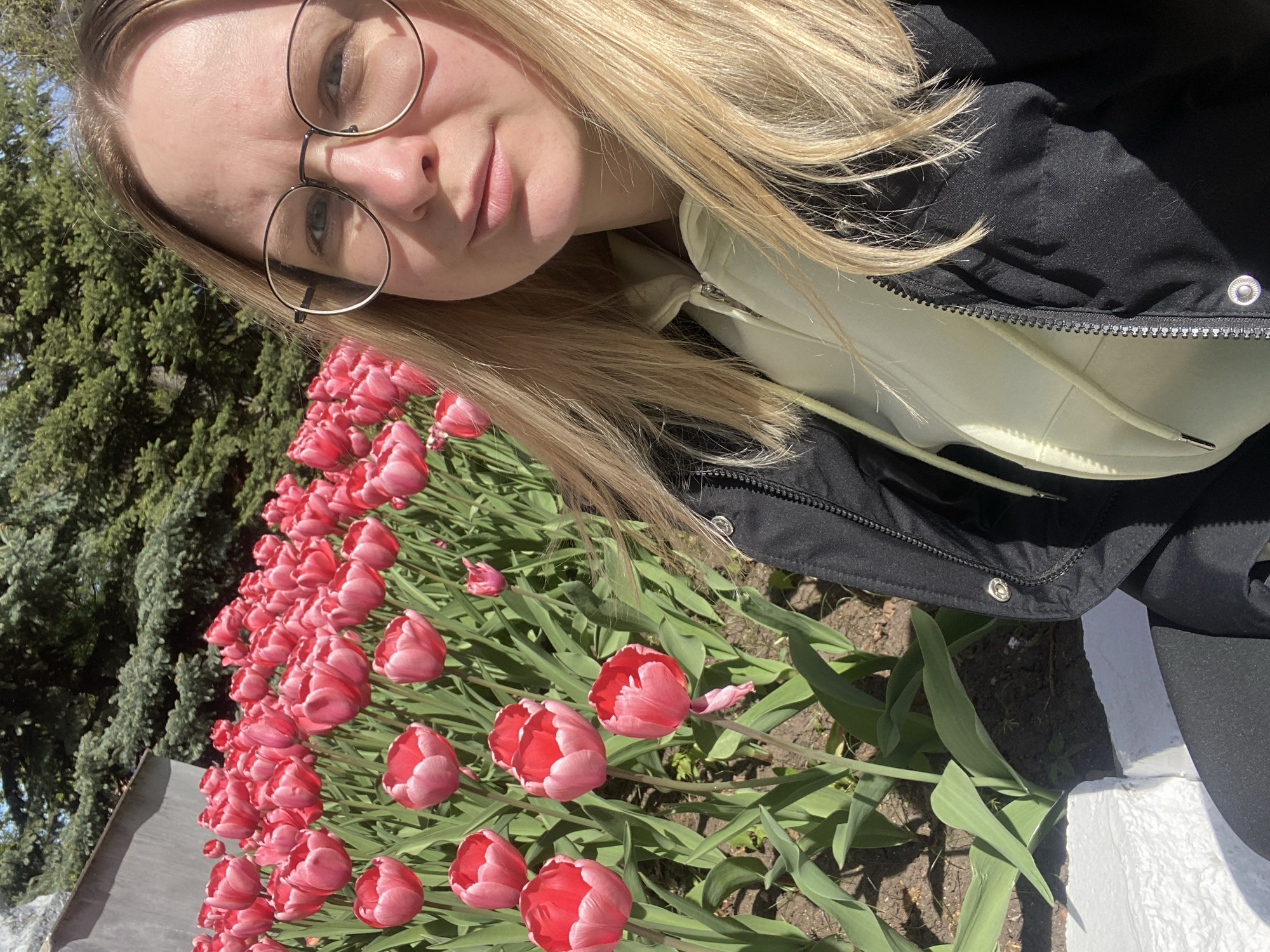 A selfie of a fair-skinned woman with blonde hair and round eyeglasses, taken in the city of Izhevsk, Russia. The woman is wearing a light-colored hoodie beneath a black jacket. She is positioned next to a flowerbed of red and pink tulips. A tree is visible in the background. A curb is visible in the bottom right corner of the image.