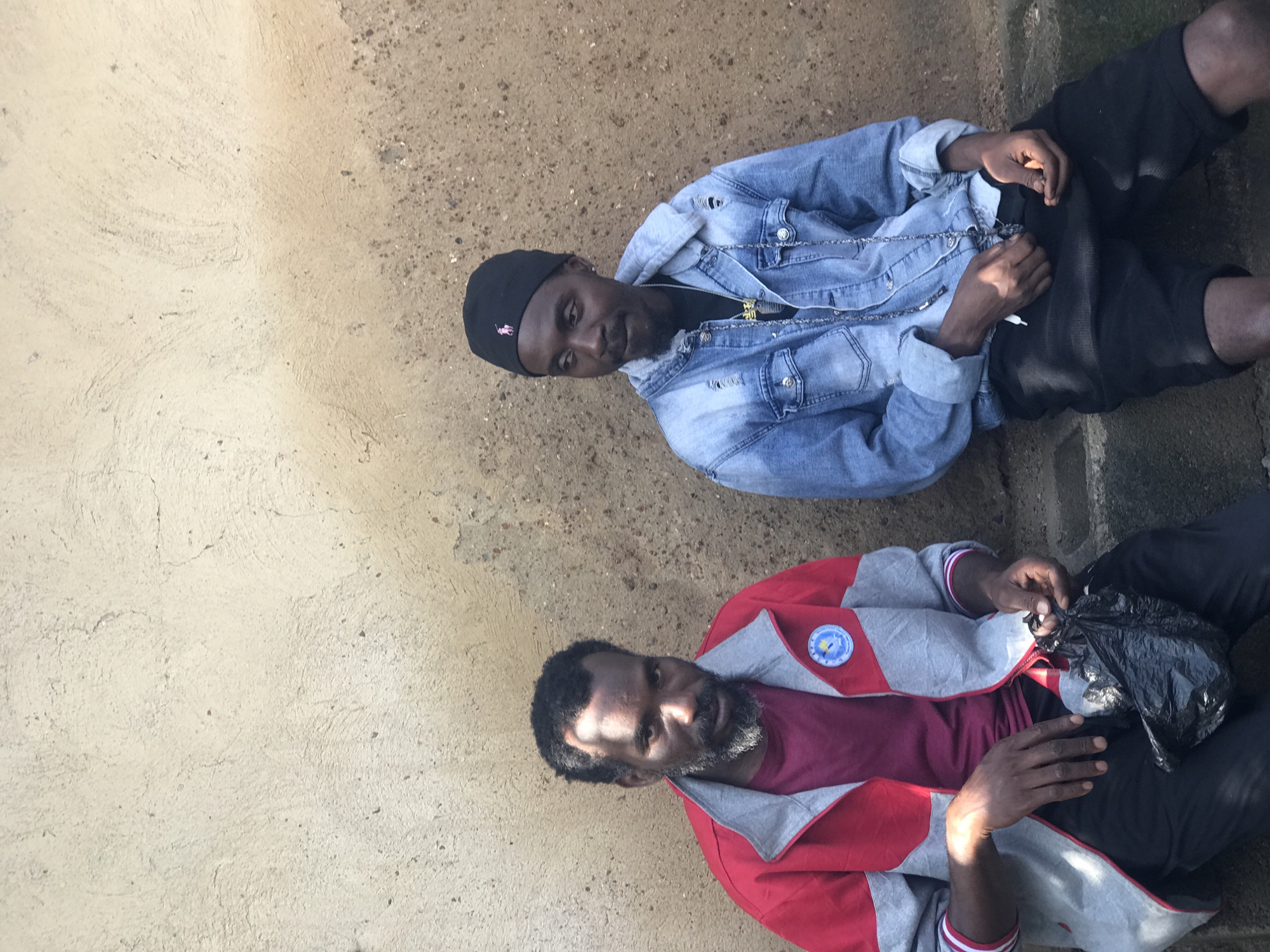Two men are sitting on a concrete step in front of a rough, beige-colored wall, likely in Kabong, Nigeria. The man on the left has dark skin, a beard, and is wearing a red and gray jacket over a maroon shirt and black pants. He is holding a black plastic bag in his hands. The man on the right has dark skin and is wearing a denim jacket over a black t-shirt, black shorts, and a black beanie with a pink logo. He is looking to his left. The wall behind them appears textured and weathered.