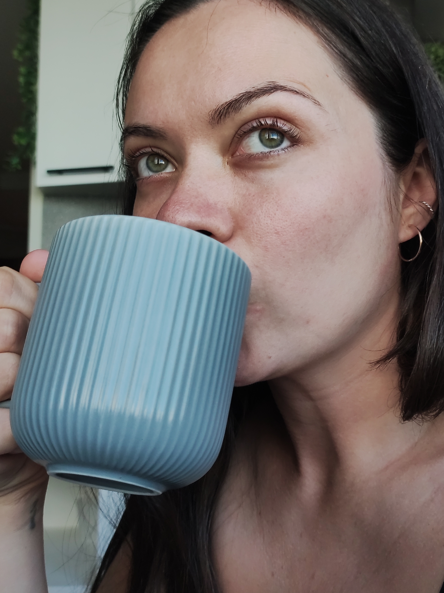 A woman with light skin, dark hair, and green eyes is drinking from a ribbed, blue mug. She is wearing a gold hoop earring and has multiple piercings in her ear. The background is out of focus, but suggests an indoor setting with a white cabinet and green foliage visible. The location of Chaikovsky, Russia is not discernible from the provided image.