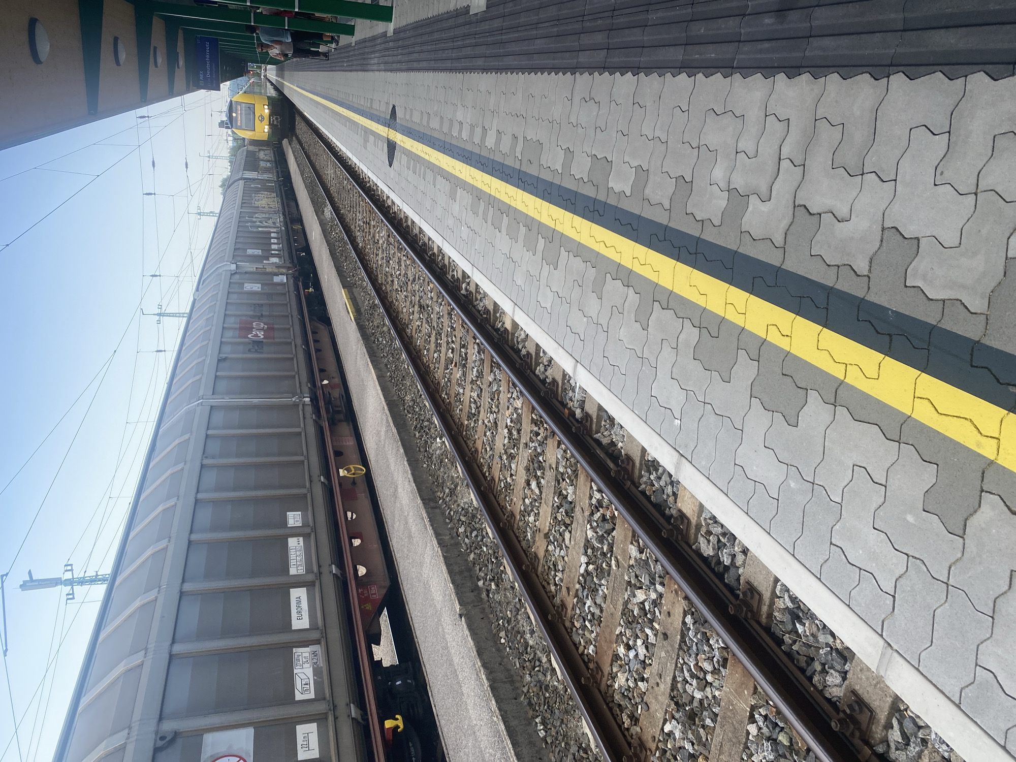 The image shows a train platform in Wulkaprodersdorf/Vulkaprodrštof, Austria. On the left, a long freight train is parked on a track. The freight cars are grey and appear to be carrying some form of cargo. The train has markings that include "EUROFIMA" and "Cargo". On the right side of the image, a yellow passenger train is approaching or stopped at the platform. Several people are visible on the platform, standing near the passenger train. The platform is paved with interlocking concrete blocks and has a yellow line marking the edge. The environment is outdoors under a clear blue sky. Overhead wires for the electric train are visible. A station roof structure extends from the right side of the frame.