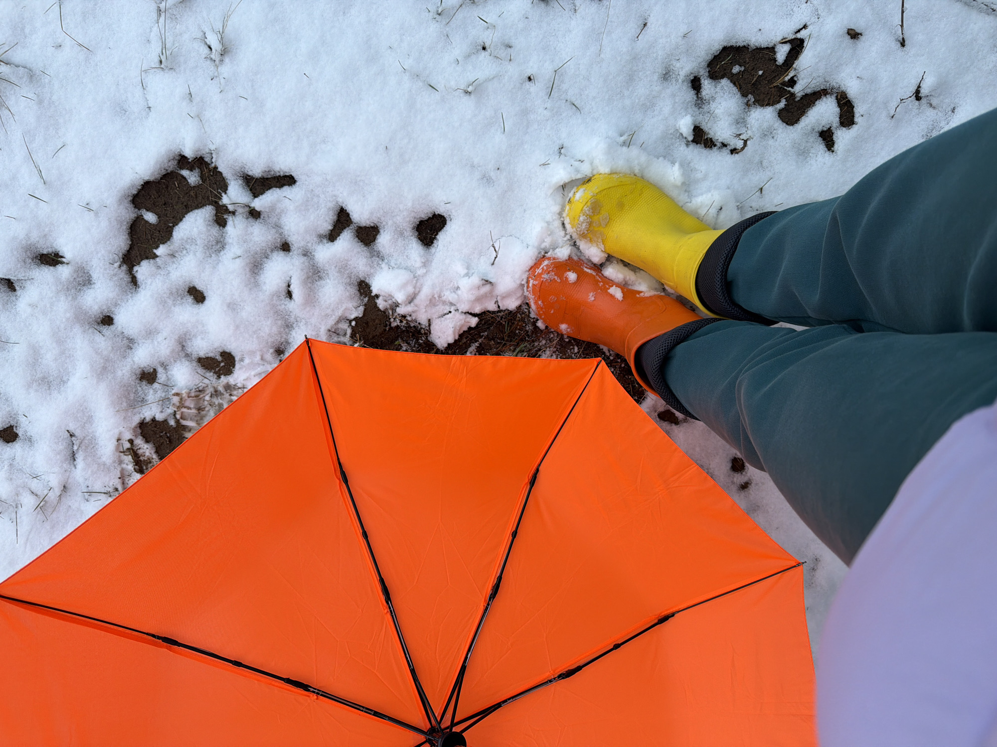 A person in orange and yellow rubber boots stands in snow with an orange umbrella. The person is wearing green pants and a white shirt. The ground is partially covered in snow. The photograph was taken outdoors, likely in a natural setting or a park.