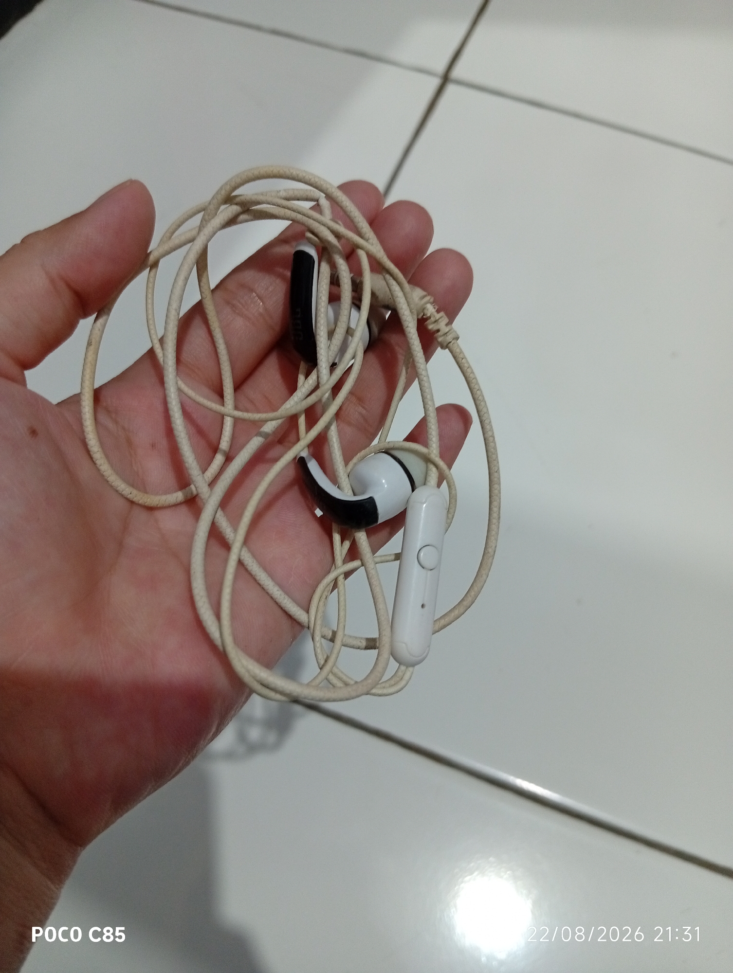 A hand holds a tangled white earbud cord with two earbuds. One earbud is partially visible, black and white, and the other is predominantly white with a black accent. A white inline microphone with a single button is attached to the cord. The background is a white tiled floor with grout lines, and a portion of a wall is visible. A timestamp in the bottom right corner reads "22/08/2026 21:31." The scene is indoors, likely a residential setting in South Tangerang, Indonesia.