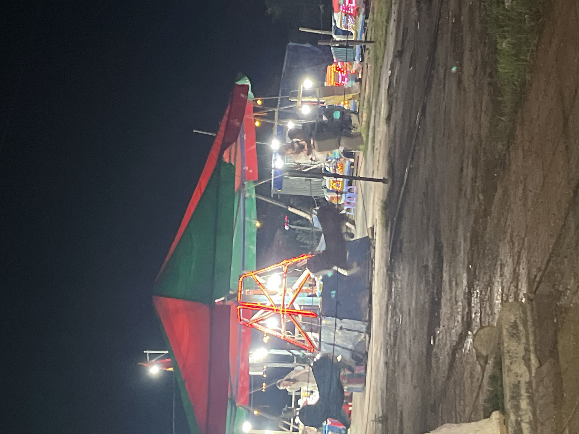 This nighttime photograph depicts a scene in Bac Nha Trang Ward, Vietnam, showcasing a local fair or market. A large tent with red and green panels dominates the left side of the frame. Inside the tent, a Ferris wheel-like ride with red supports is illuminated. People are visible inside and around the tent. Further back, stalls and other attractions, also lit up, stretch along what appears to be a road or pathway. The ground surface to the right appears uneven and perhaps muddy. The night sky is dark.