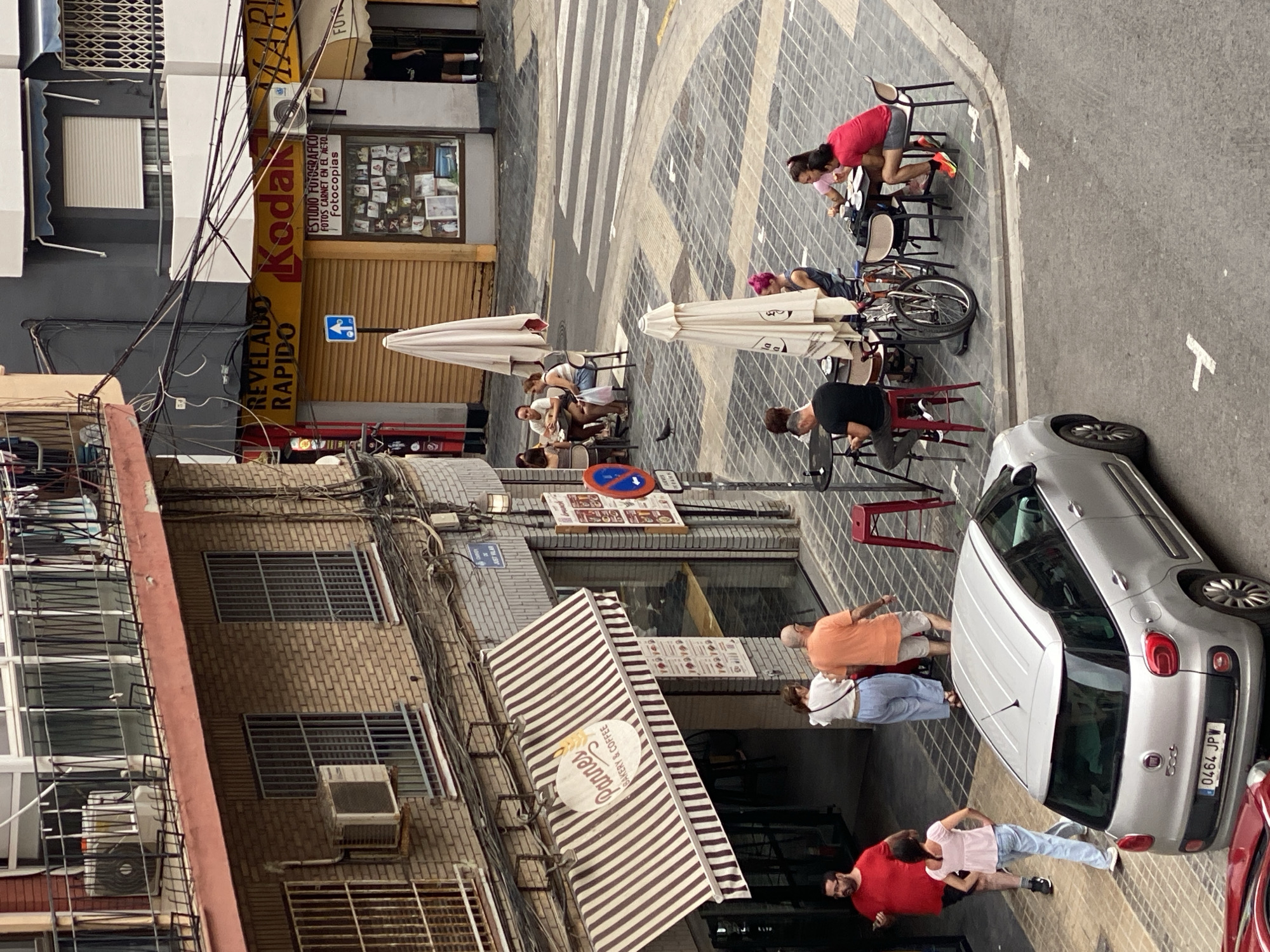 The image captures a street scene in Valencia, Spain, focusing on the intersection of a narrow street and a wider road with a crosswalk. On the left, a building with a brick facade features a bakery and coffee shop called "Pannes." An awning with brown and white stripes shades the entrance, where several patrons are visible. A silver Fiat 500 car is parked on the street in the foreground.
Across the street, a building with a yellow awning displays "Revelado Rapido" and "Kodak." Adjacent to this, a store with a window display showcases photographs and has a sign for "Estudio Fotografico." Several tables with umbrellas are set up on the sidewalk, with groups of people sitting and interacting. One person is seen sitting on a red stool at a table. A bicycle is parked near one of the tables.
The ground is paved with patterned tiles, and white lines mark the crosswalk. A pigeon is observed on the tiled ground. The overall atmosphere suggests a casual, urban setting with ongoing commercial activity and people enjoying outdoor seating. The buildings exhibit a mix of architectural styles, common in European cities.