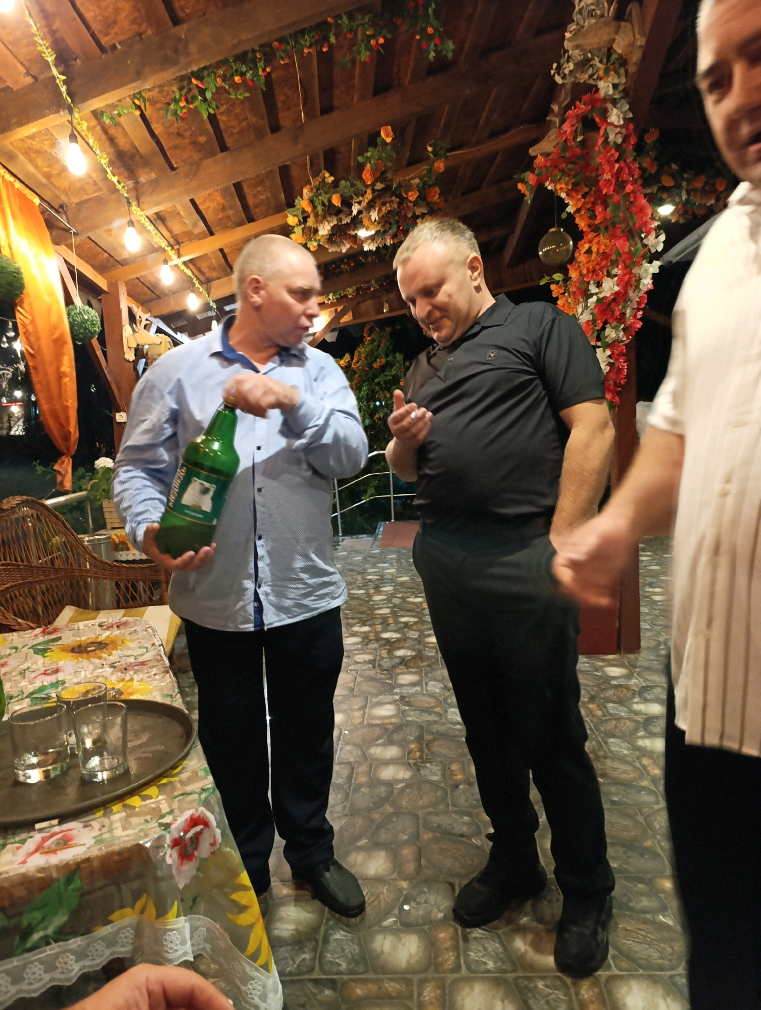 In Korzhevo, Moldova, three men are seen in an outdoor, covered patio area. The man on the left, wearing a light blue button-down shirt and dark pants, holds a large green bottle. The man in the middle, dressed in a black polo shirt and black pants, gestures with his right hand. A third man, partially visible on the right, wears a white striped shirt. The patio features wooden beams, hanging decorative flowers, and string lights. A table covered with a floral tablecloth is set with glasses and a tray. The ground is paved with stones.
