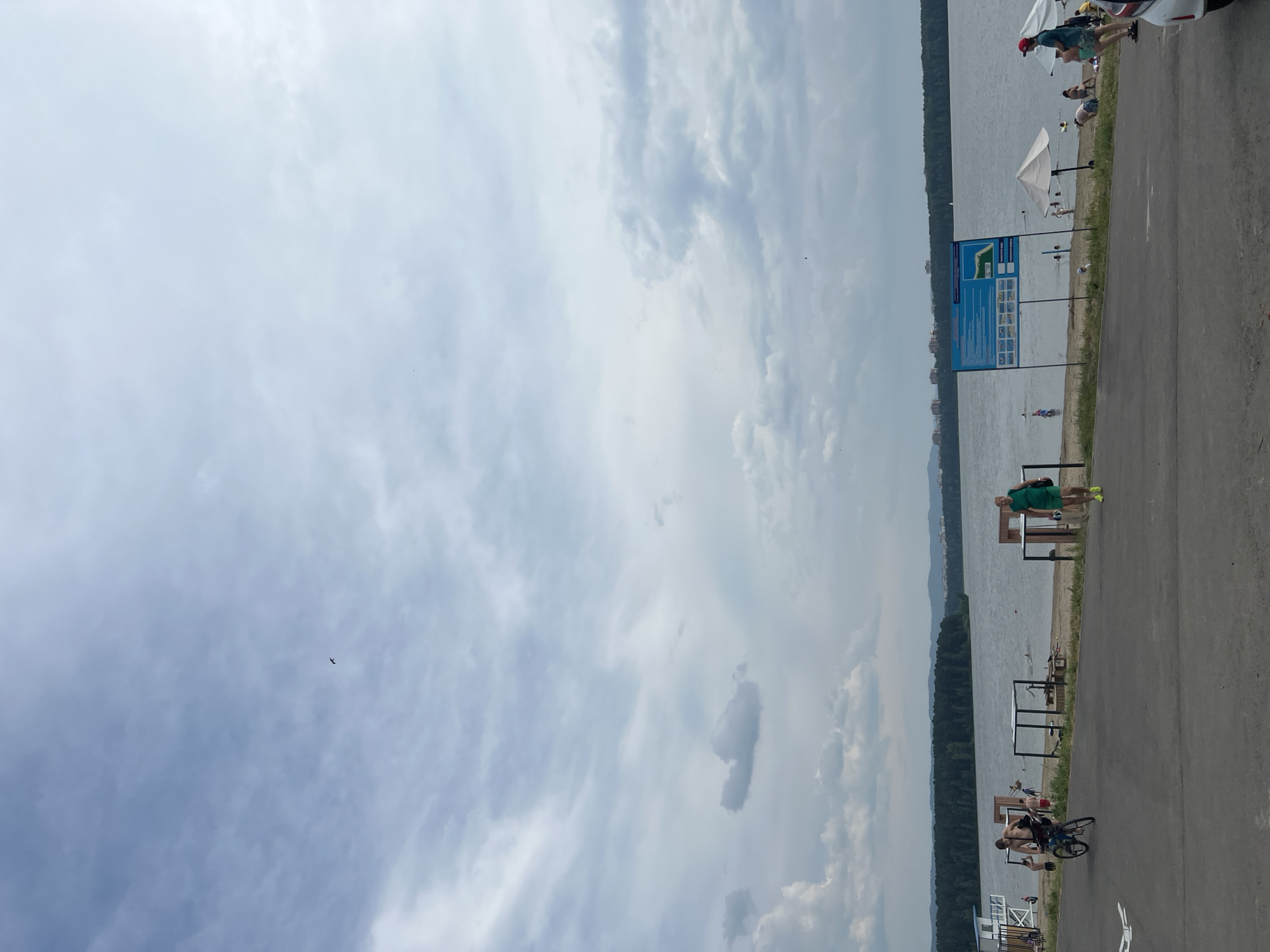 This media file depicts a scene at a waterfront location, likely a beach or recreational area, in Zheleznogorsk, Russia. The image is oriented vertically, with the sky occupying the left two-thirds and the waterfront and surrounding area on the right. The sky is filled with a mix of white and gray clouds, suggesting overcast or partly cloudy weather. A small airplane is visible in the upper portion of the sky.
The waterfront features a body of water, possibly a lake or river, bordered by a tree-lined shore in the distance. Along the edge of the water, there is a paved path or road. Scattered along this path are several people engaged in various activities. In the foreground on the right, a person is riding a bicycle. Further along the path, near some tables and what appear to be sunshades, are other individuals. One person is sitting on a bench, while others are visible in or near the water. A blue informational sign with diagrams and text is prominently displayed near the water's edge.
The time of day appears to be daytime, given the lighting. The overall atmosphere seems relaxed, indicative of a recreational outing. There are no strong emotional cues evident. The presence of a beach umbrella, people in casual attire, and the waterfront setting suggest a leisurely summer day. The text on the informational sign is not legible enough to be transcribed.