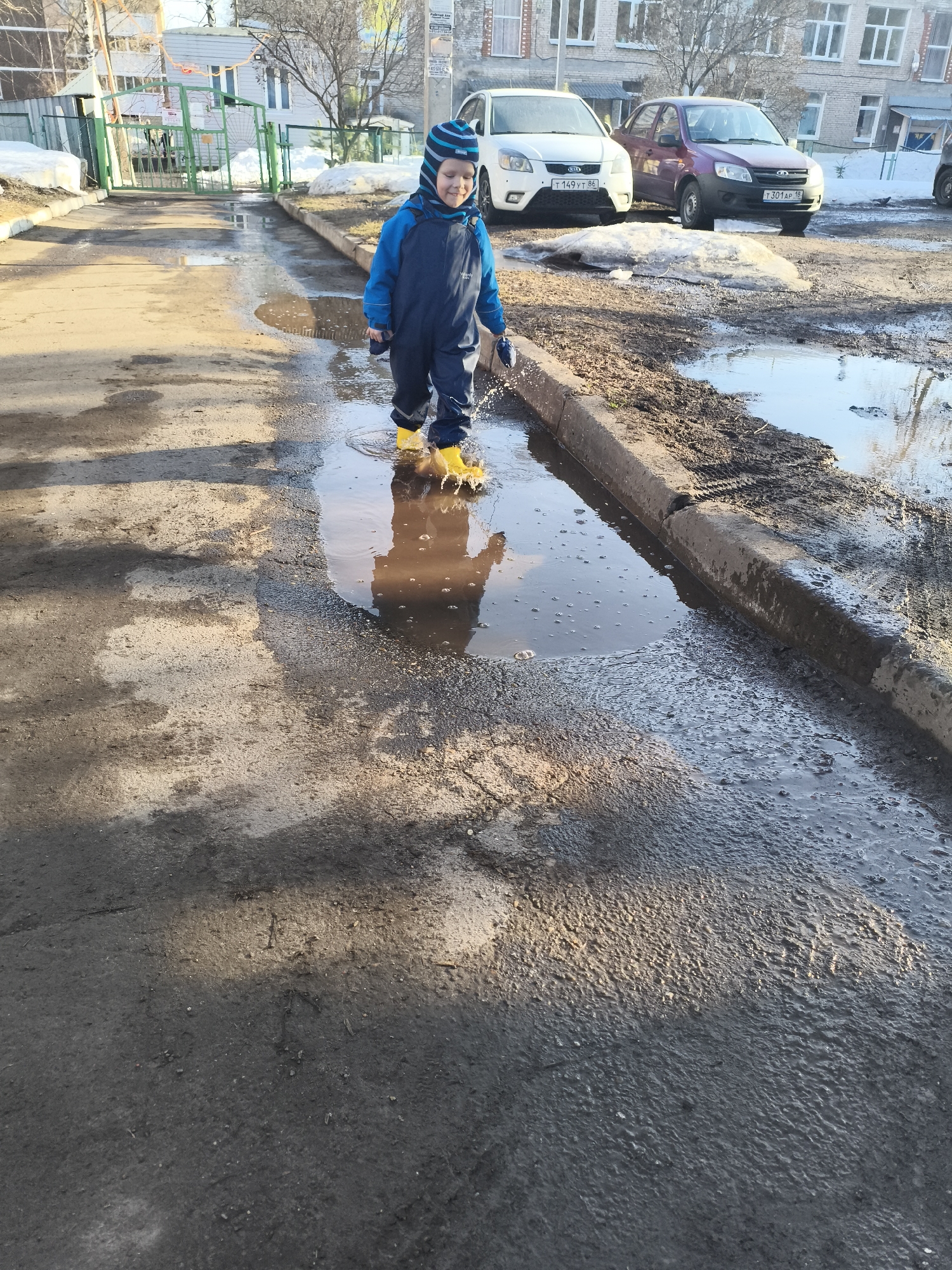 A young child in a blue snowsuit and yellow rubber boots is splashing in a large puddle on a paved surface. The child is wearing a blue and white striped hat and gloves. The puddle reflects the child's image. The surrounding area is muddy and appears to be a parking lot or street. In the background, there are several cars parked near a building with multiple windows. There is also a green gate visible. Based on the license plates of the cars in the background (T 149 YT 86 and T 301 AP 18), this image was taken in or around the city of Izhevsk, Russia. The presence of melting snow and bare trees indicates that this photograph was taken in early spring.