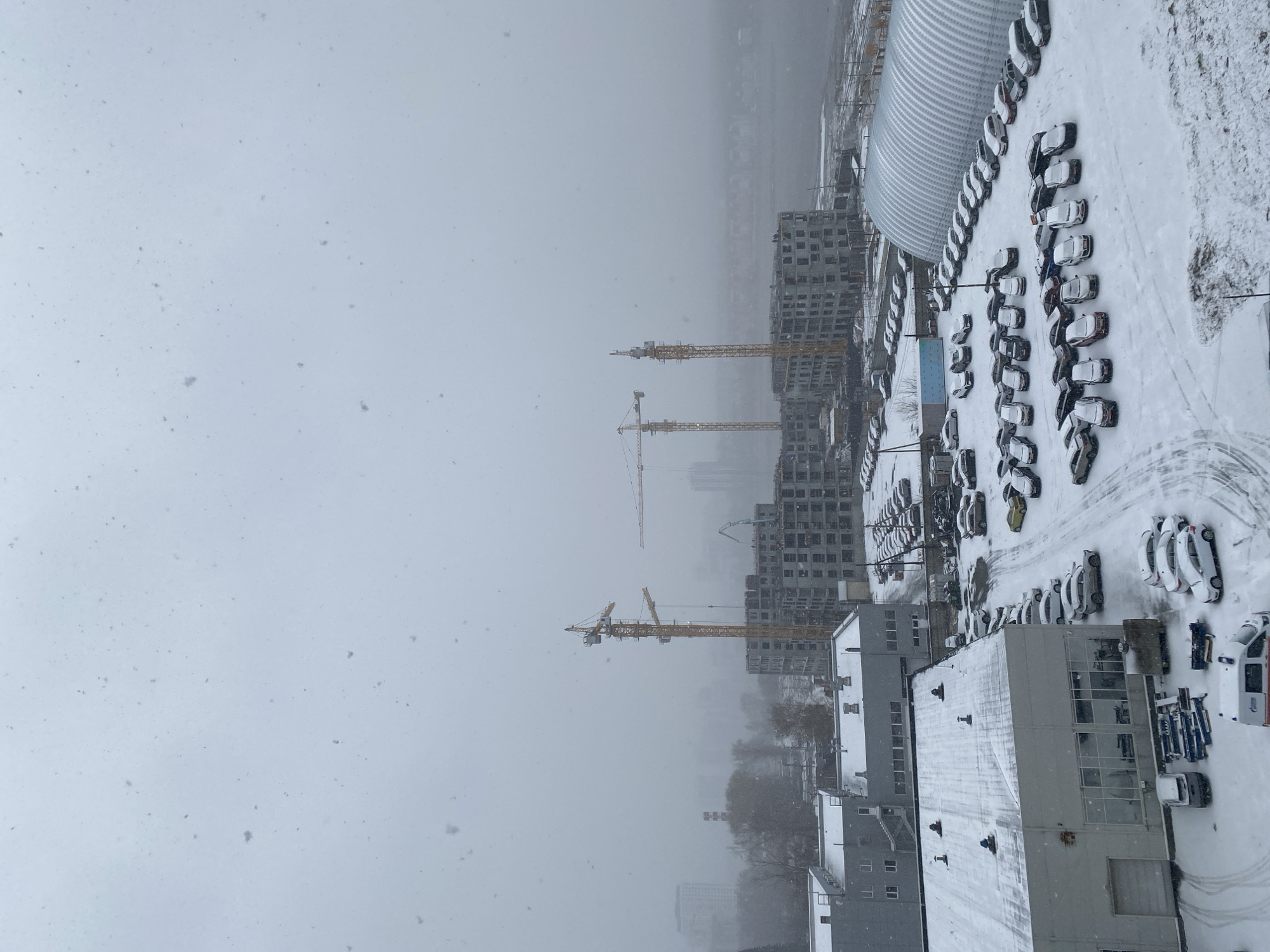 The image depicts a snow-covered urban landscape in Yekaterinburg, Russia. The scene appears to be taken from an elevated vantage point, looking down on a construction site with several unfinished high-rise buildings and cranes. Snow is falling heavily, obscuring the view. A parking lot is filled with snow-covered cars. The photo includes buildings, construction cranes, and cars covered in snow.