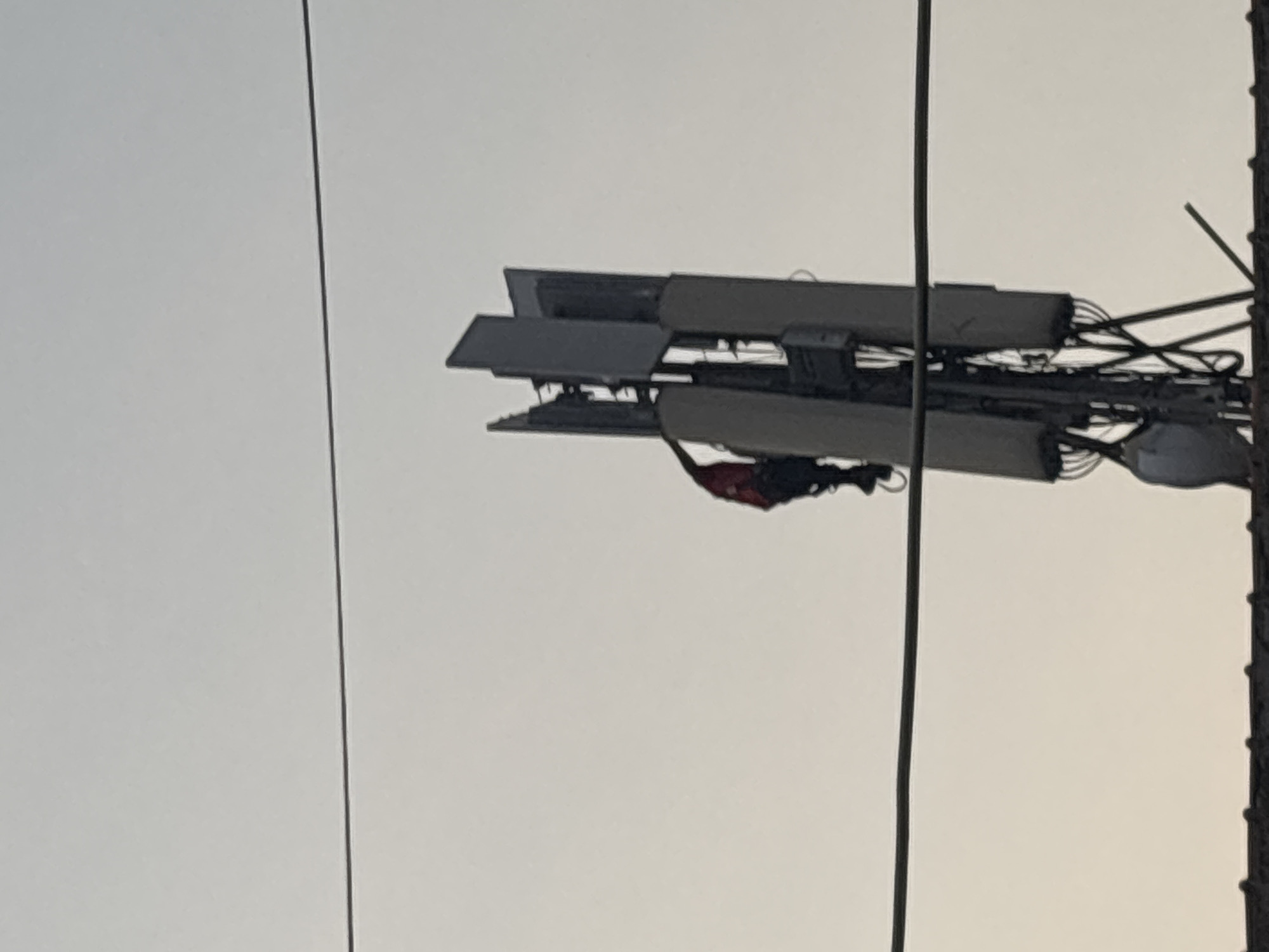 The image shows a telecommunications tower in North Jakarta, Indonesia. The tower is equipped with multiple cellular antennas. Utility cables are visible in the foreground. A piece of fabric is draped on the antenna. The sky is overcast.