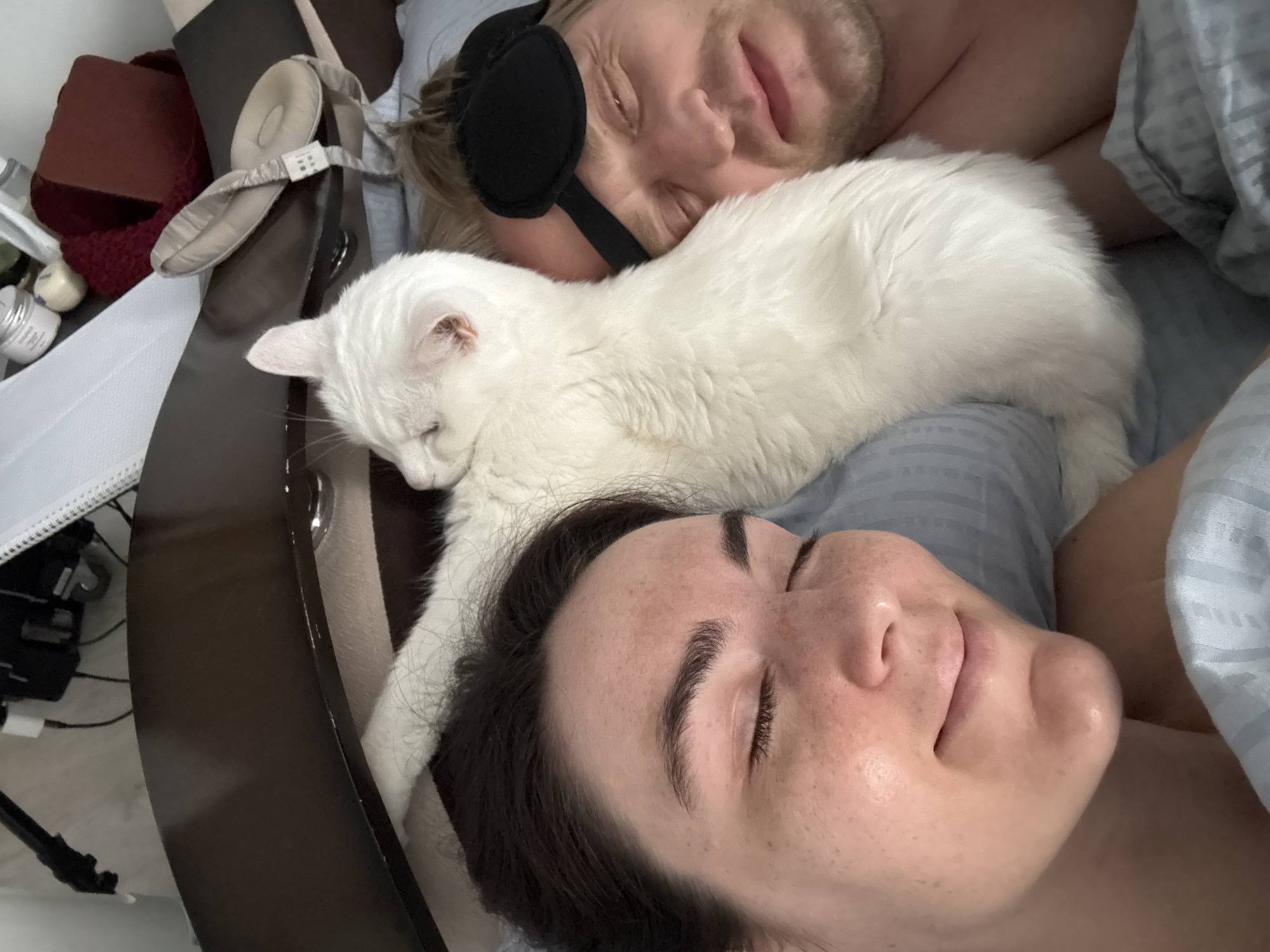 A close-up, eye-level shot captures two sleeping individuals and a white cat in a bed. The woman, with dark hair, lies with her head near the bottom of the frame and her face turned towards the camera. To her left is a man with blonde hair wearing a black sleep mask. A white cat rests on top of them. The bed is covered with blue and grey bedding. In the background, on the left, are various objects, including a maroon-colored item, skincare products, and what appears to be a sleep-aid device on a dark brown surface. There is no clear evidence to determine if this photograph was taken in Tyumen, Russia.