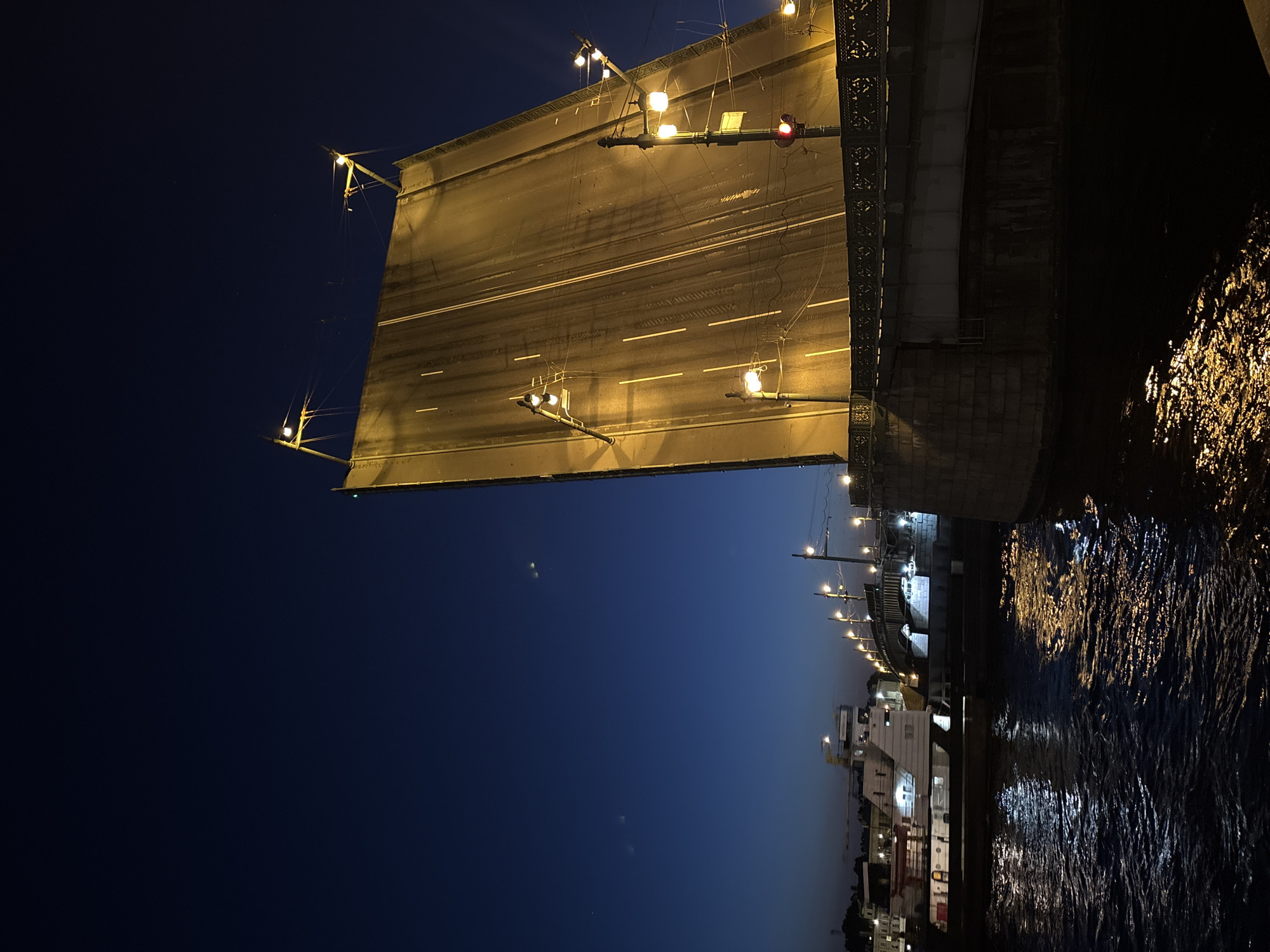 The image shows a drawbridge at night in an unknown city. The bridge deck is raised, with traffic markings visible under the artificial lighting. A red signal light is attached to the bridge's mechanism. Dark water reflecting light is visible below and to the right of the bridge. A building is to the right of the bridge, alongside a line of street lights. The night sky transitions from dark blue to light blue.