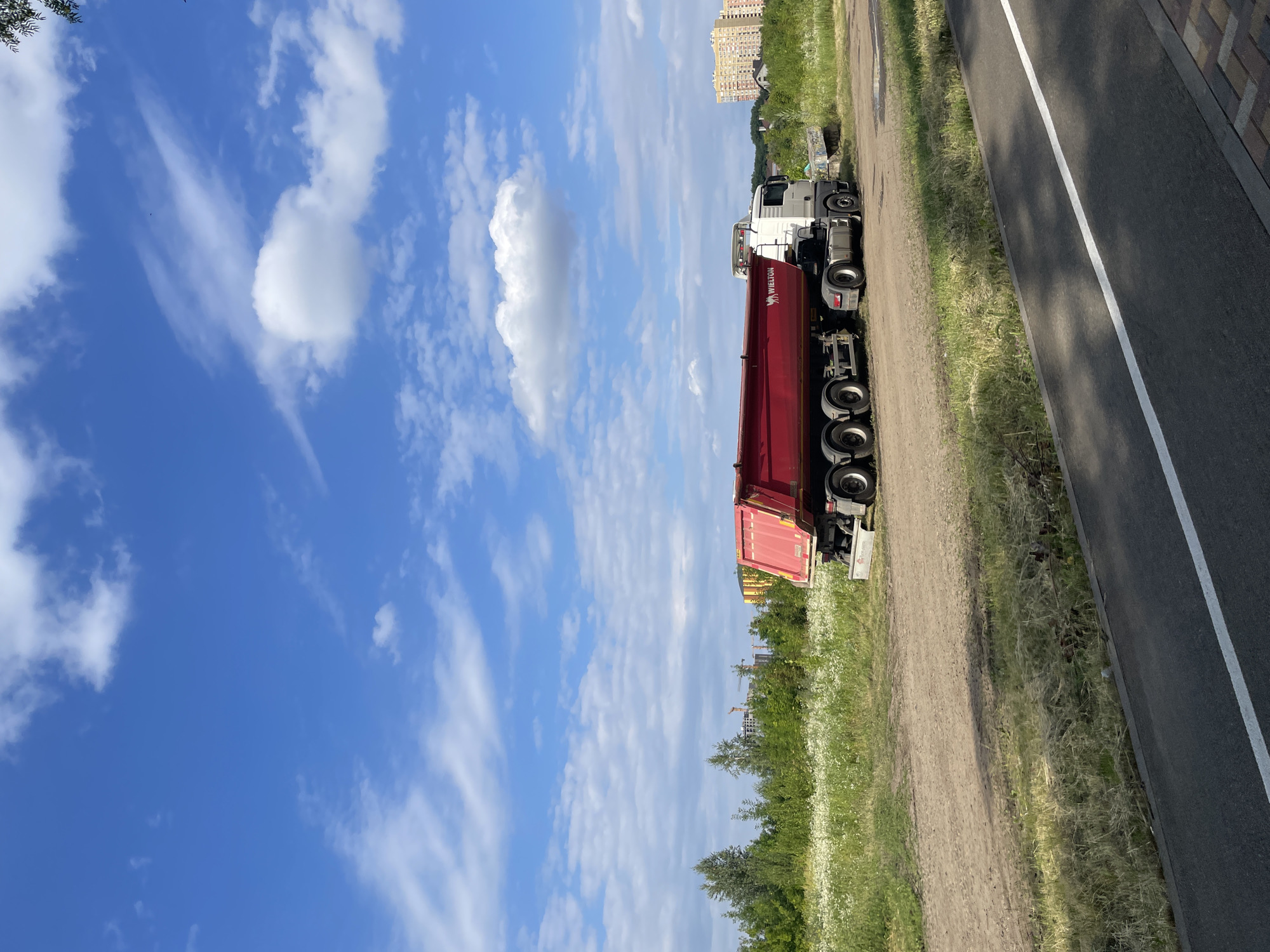A red dump truck with the "WIELTON" logo is parked on a dirt patch next to a grassy field under a blue sky with scattered white clouds. In the background, there is a dense line of green trees, construction cranes, and several multi-story buildings. In the foreground, a paved path with a white line runs parallel to the grassy field. The image does not depict the city of Stavropol, Russia; it appears to be an outdoor construction or industrial area. There are no people visible in the scene.