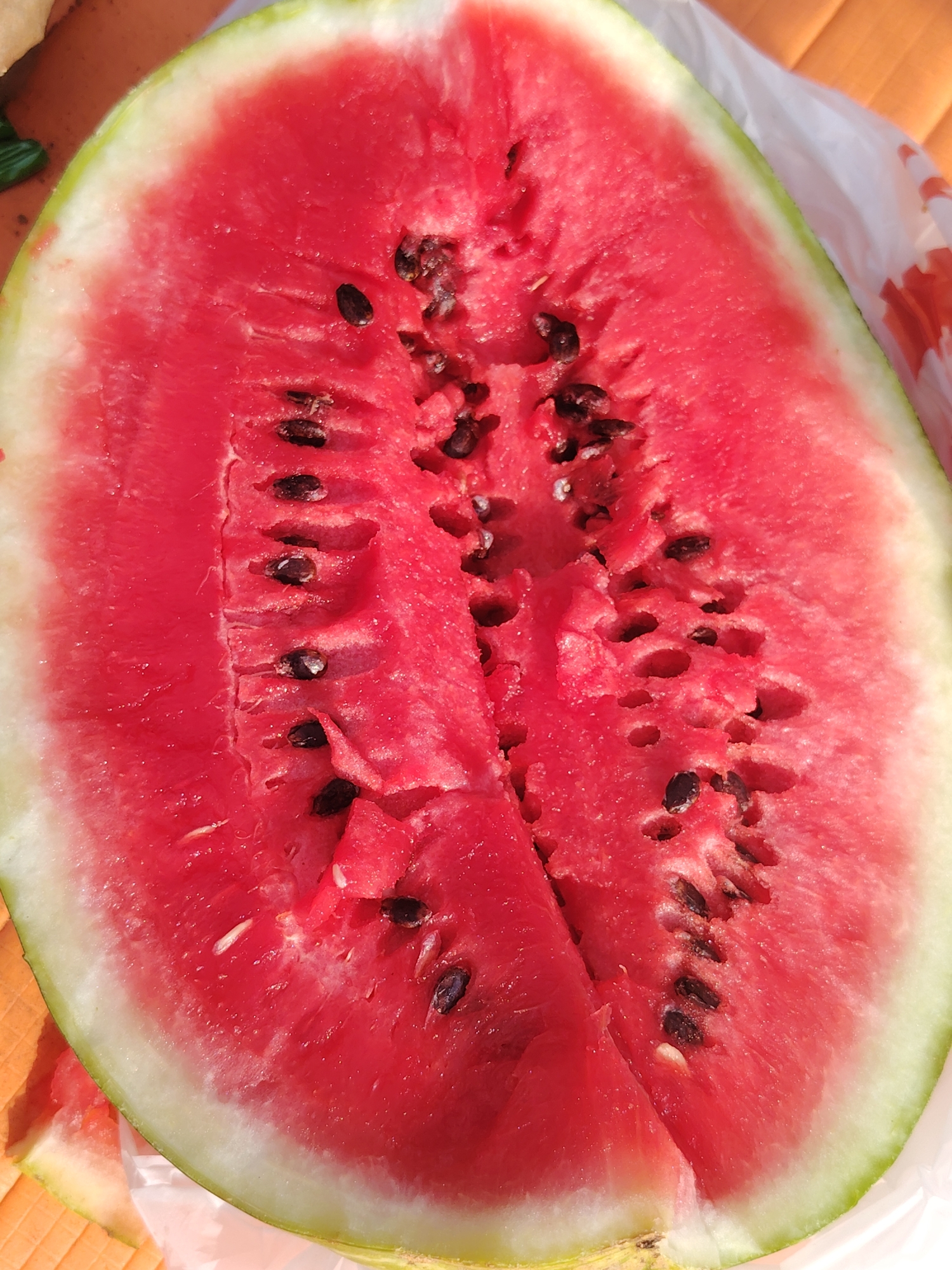 A close-up, overhead shot shows a halved watermelon resting on a surface that appears to be a wooden table. The watermelon flesh is bright red and juicy, with a scattering of black seeds visible. The rind is green and white. A piece of white plastic wrap is visible on the right side of the frame. The overall setting suggests an outdoor or casual environment, possibly a picnic or a market stall in a city like Perm (part of the Perm Krai, formerly known as Прикамский region in Russia).