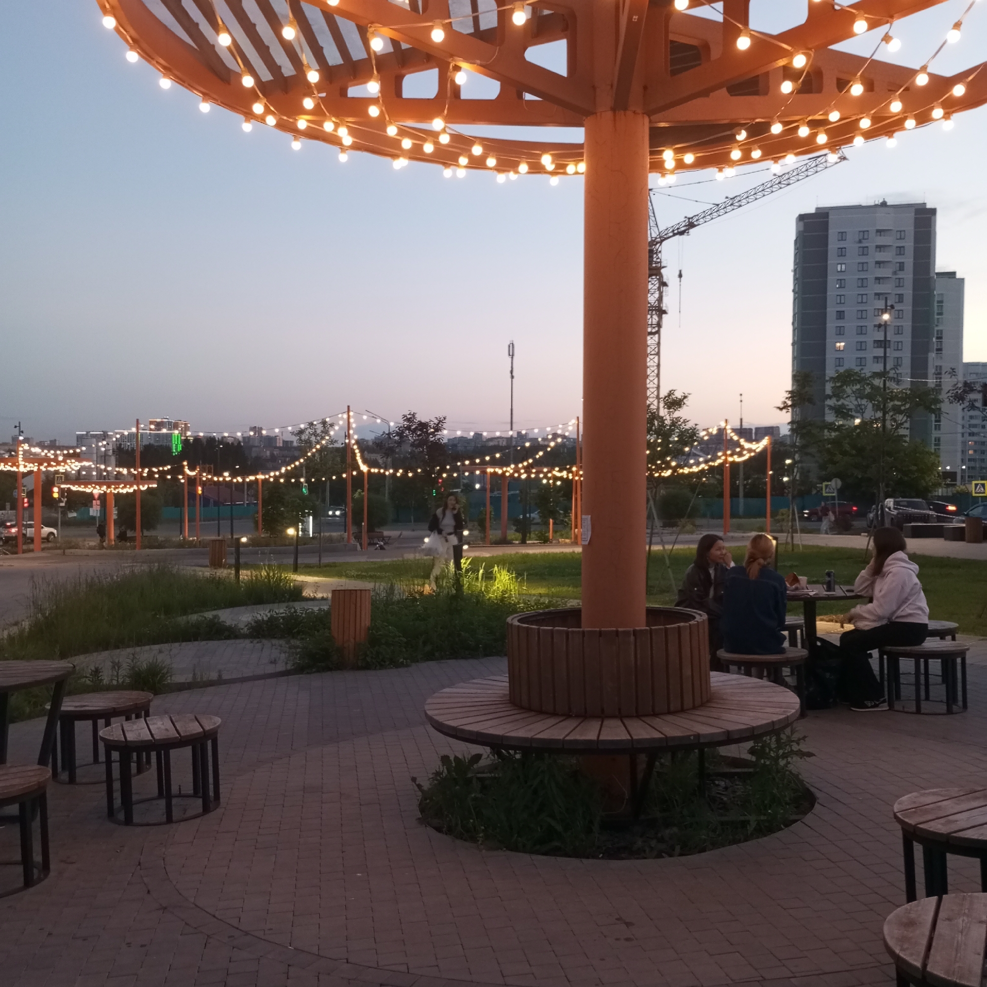 The scene depicts an outdoor urban public space in the city of Pervomaysky, Russia, at dusk or early evening. The sky is clear, transitioning from a light blue horizon to a darker, deep blue zenith. Artificial lighting is active throughout the area.
A large, circular, open-framework wooden canopy structure, adorned with numerous illuminated warm white spherical string lights, is centrally featured. This structure is supported by a prominent, cylindrical, light brown central pillar. Around the base of this pillar, a circular wooden bench is integrated.
To the right of the central pillar, three young adult females are seated at a round wooden table, each on a separate wooden stool. The female on the far right wears a light-colored hoodie and dark pants, with dark hair. The female in the middle has reddish-blonde hair and wears a dark blue top. The female on the left wears a dark jacket and has dark hair. They appear to be engaged in conversation, with cups or bottles visible on their table.
In the mid-ground, to the left of the central pillar, a single young adult female is walking on a paved pathway. She is dressed in a dark jacket over a light-colored top and skirt, and is carrying a light-colored bag.
The foreground ground cover consists of reddish-brown interlocking pavers, interspersed with patches of green grass and low-lying vegetation around the furniture and pillar base. Additional wooden stools and a table are partially visible in the far left foreground.
The background reveals a multi-lane road with faint vehicular traffic. A green traffic light is illuminated on the left. Numerous strings of warm white spherical lights are strung between orange-colored poles, extending into the distant background. Several multi-story, modern apartment buildings, predominantly grey-blue with grid-patterned windows, are visible on the right. A large construction crane is present in the mid-background, to the left of the prominent apartment building.