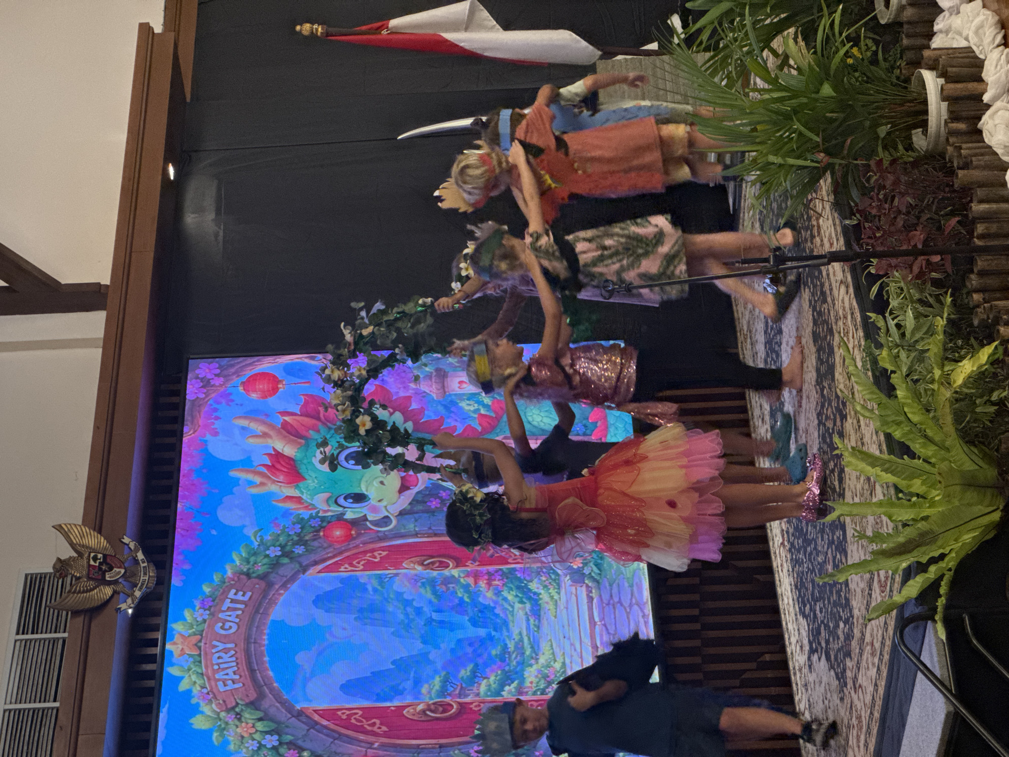 A group of children, dressed in costumes, are performing on a stage in front of a large digital screen displaying a "Fairy Gate" scene with a cartoon dragon. The children are wearing crowns, floral headpieces, and colorful dresses. Some are holding greenery. The stage has a patterned rug and is decorated with potted plants. In the background, a flag of Indonesia is visible. The overall scene suggests a theatrical performance or event for children. The location is the city of Kuta Selatan, Indonesia.