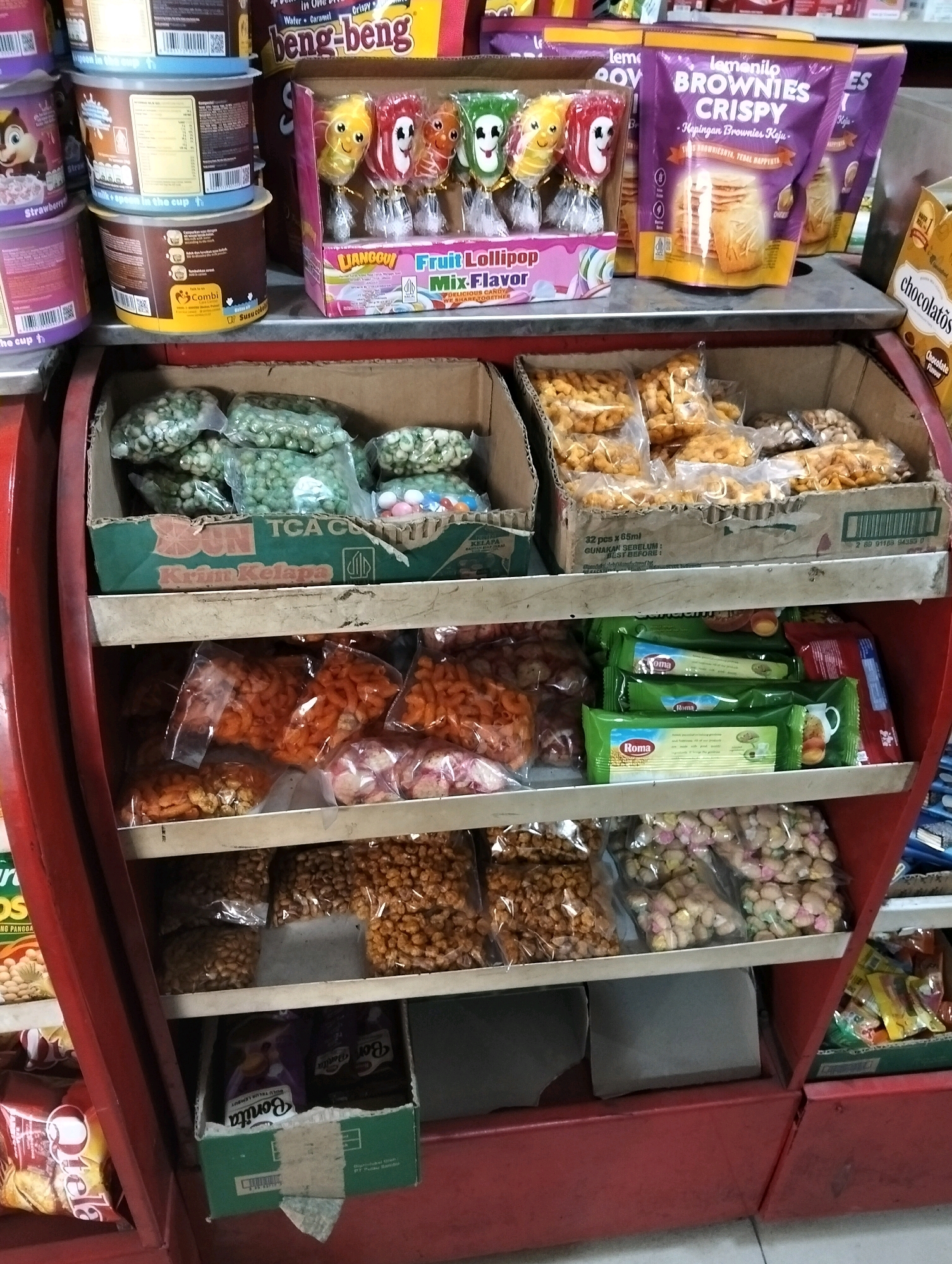 The image depicts a retail display of various packaged snacks and candies within a store. The display consists of multiple shelves, with items neatly arranged in their original packaging. On the top shelf, there are several boxes of what appear to be biscuits or cookies, some with prominent branding like "Beng-beng" and "Brownies Crispy". To the left of these, colorful lollipop candies are visible, individually wrapped and arranged in a display box with text that includes "Liancouy Fruit Lollipop Mix Flavor".
The second shelf from the top holds large, clear plastic bags filled with green-colored snacks, likely peas or beans. Beneath these are boxes containing more packaged snacks, some appearing to be a savory, crunchy variety in orange and brown hues.
The third shelf from the top features several green-packaged snack items, with the brand name "Roma" visible on some of them. Next to these, more plastic bags filled with snack items are present.
The bottom shelf displays more plastic bags of various snacks, including some with a reddish-pink hue and others that are a light beige color. In the lower right corner, a green box with colorful wrappers is visible, containing more snack items. The lower portion of the display unit is red. The overall environment suggests a small shop or convenience store. The text "Cileungsi, Indonesia" is not explicitly visible in the image itself but serves as the location context for this scene.
