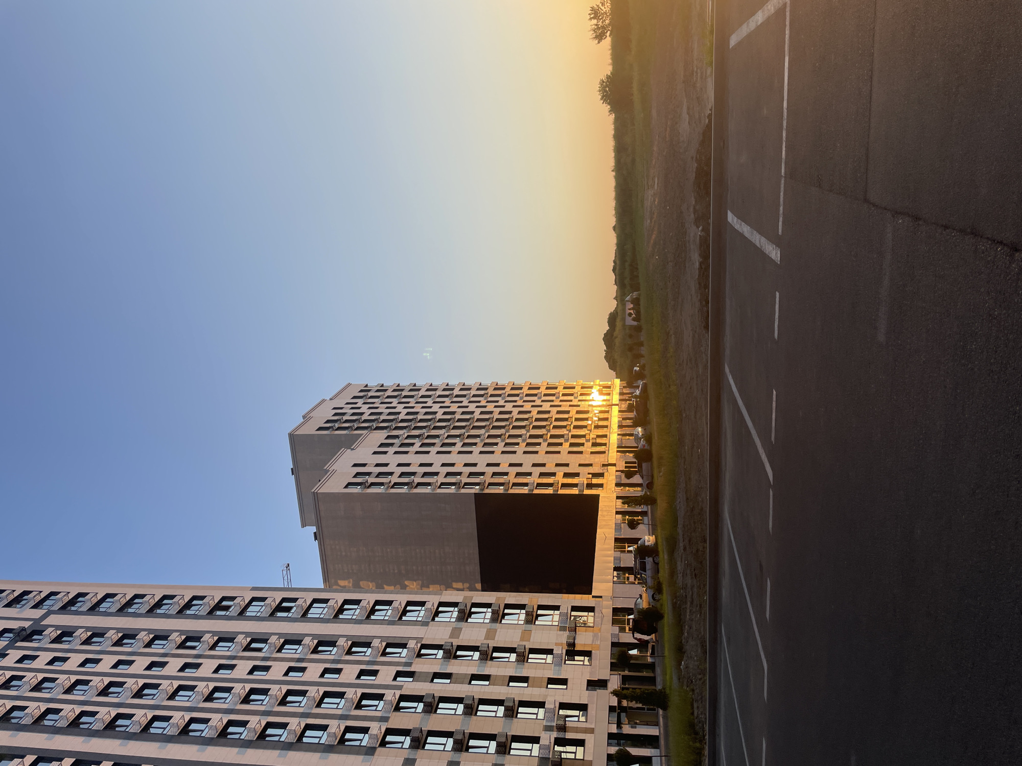 An exterior shot captures two modern high-rise buildings under a clear sky during sunset in the city of Stavropol, Russia. The building on the left, mostly out of frame, is light-colored with a grid of windows and dark accents. To its right stands a taller, similarly designed building, its facade illuminated by the warm glow of the setting sun. A large, dark rectangular section is visible on the side of this building, possibly a screen or a darkened facade.
In the foreground, an asphalt parking lot is marked with white lines indicating parking spaces. The pavement shows some signs of wear. Behind the parking lot, a strip of green grass and some trees separate it from the buildings. Beyond the buildings, a field of dry grass stretches towards the horizon, where the sun is setting, casting a golden light. A few parked cars are visible near the entrance of the buildings, and a small group of people are standing near them. The overall scene depicts a contemporary urban landscape at dusk.