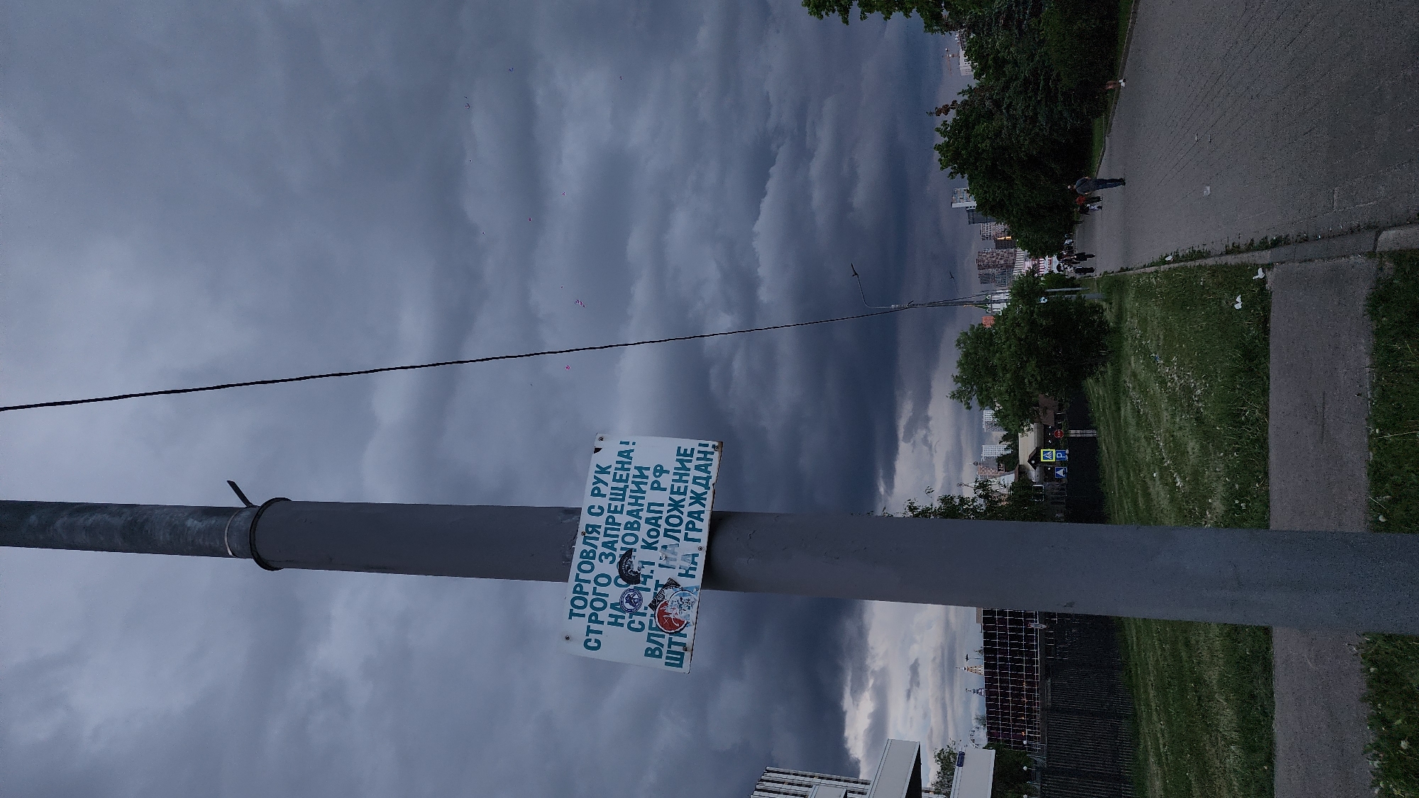 The image shows a cloudy day in what appears to be a public space in Moscow, Russia. A metal pole stands in the foreground with a sign attached that reads, "ТОРГОВЛЯ С РУК СТРОГО ЗАПРЕЩЕНА! НА ОНОВАНИИ СТ 14.1 КоАП РФ ГОСТ НАЛОЖЕНИЕ НА ГРАЖДАН!" which translates to "Trading by hand is strictly prohibited! Based on Article 14.1 of the Code of Administrative Offenses of the Russian Federation GOST Imposition on citizens!". In the background, there is a sidewalk with people walking, trees, and buildings. A power line runs diagonally across the sky.
