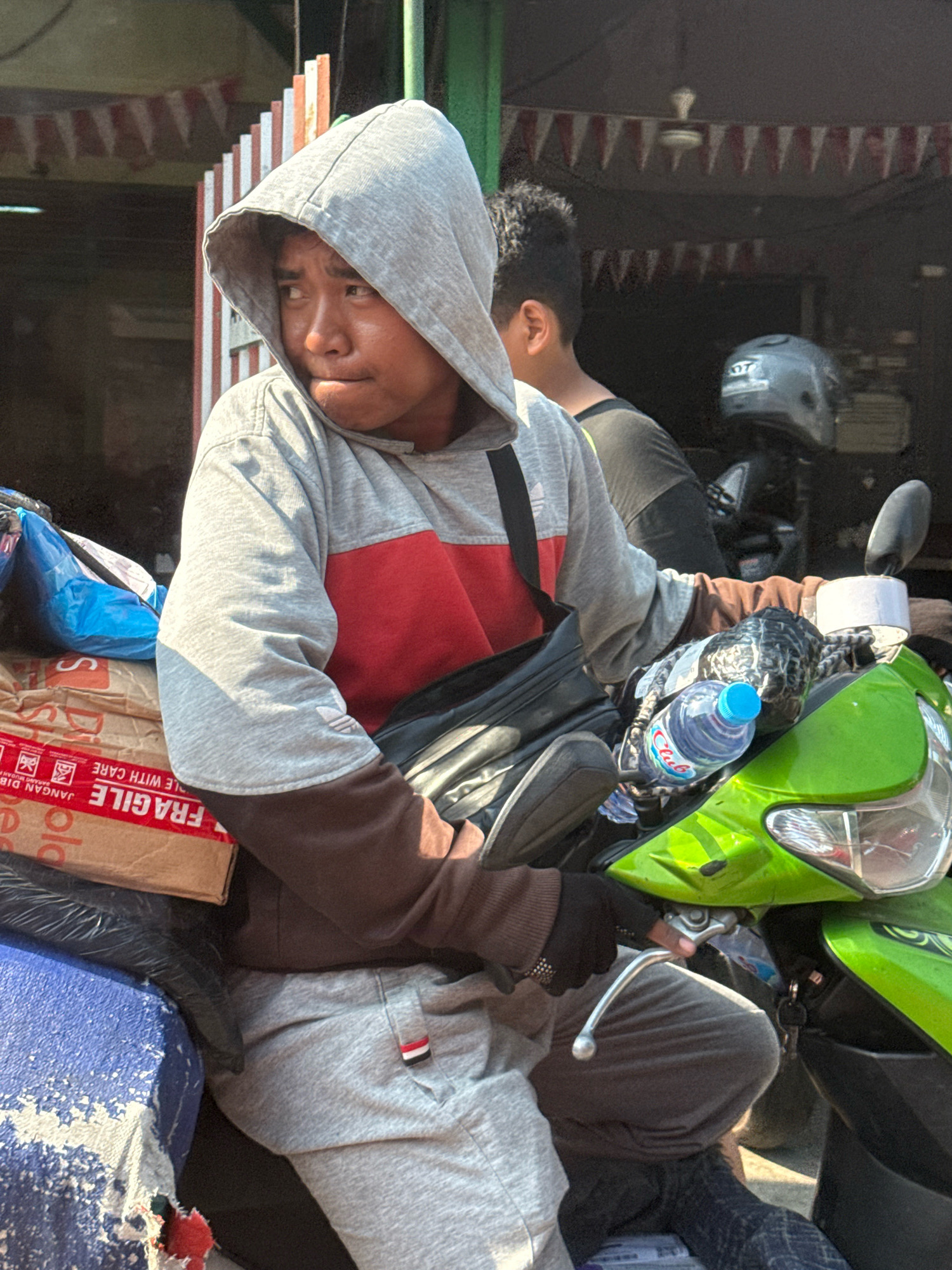 The image captures a young male, possibly a delivery person, sitting on a bright green scooter in what appears to be a commercial area of Surabaya, Indonesia. He is wearing a grey hooded sweatshirt with a red panel on the chest and brown sleeves, along with grey sweatpants. He has a black crossbody bag slung over his shoulder and is wearing black gloves. His gaze is directed to his left, with his mouth slightly open as if in thought or mid-conversation.
The scooter is laden with packages. Notably, a brown cardboard box with red "FRAGILE" and "JANGAN DIBANTING" (Do Not Slam) markings is prominently displayed. A blue plastic bag is also visible amongst the cargo. A bottle of "Club" mineral water rests on the scooter's front.
In the background, another person with curly dark hair is partially visible, wearing a dark t-shirt. Behind them, a building with red and white pennant banners strung across the entrance suggests a festive or communal atmosphere, typical of local shops or events in Indonesia. A ceiling fan is visible inside the building. A grey motorcycle helmet sits on a shelf or counter within the shop. The overall lighting suggests it is daytime, with harsh shadows indicating bright sunlight. The environment appears to be an outdoor street scene in a bustling part of Surabaya, likely a commercial district given the shops and activity.